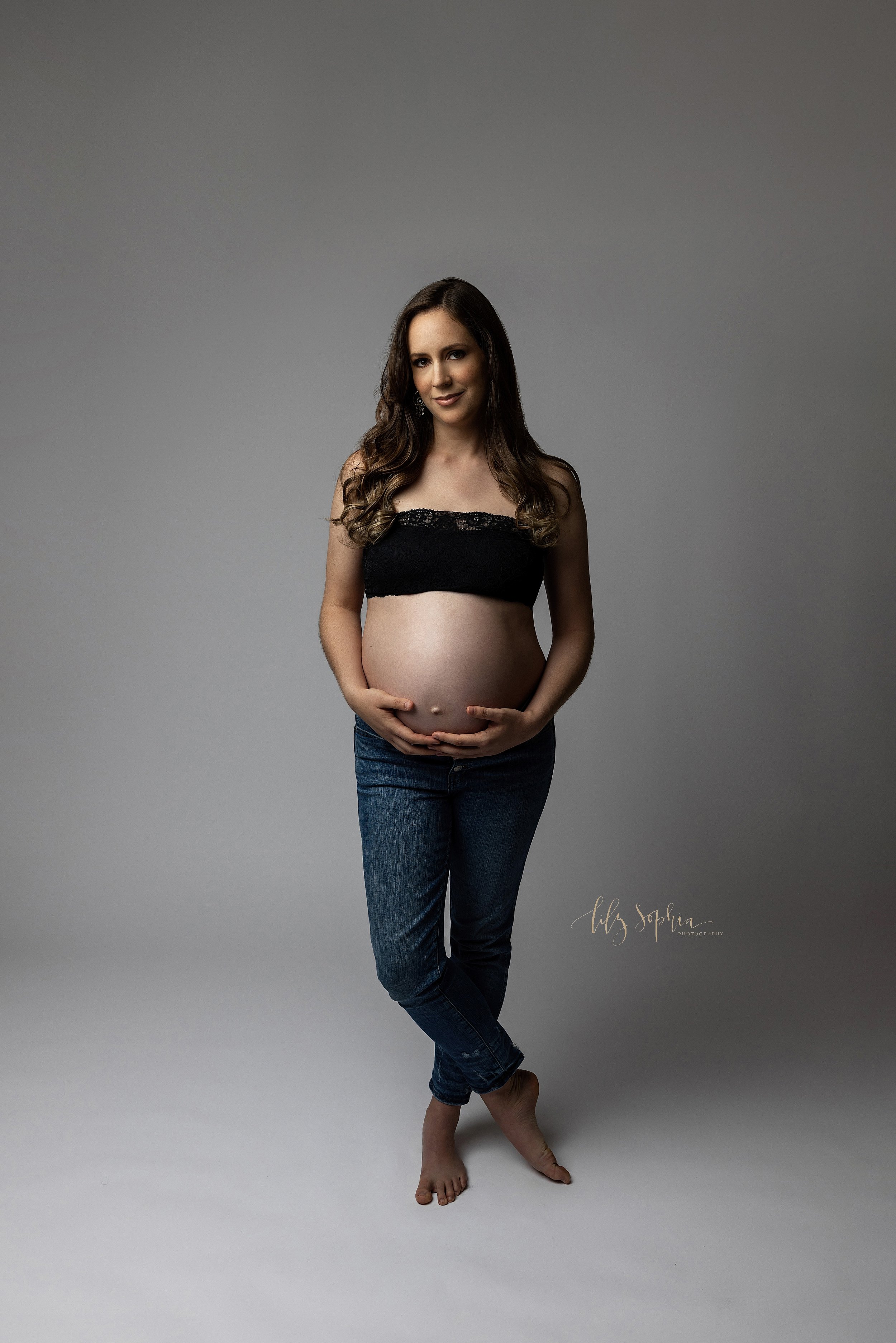  Modern maternity photo of a pregnant woman as she stands in a natural light studio wearing a black bandeau bra and a pair of button-downed blue jeans while holding the base of her belly with her hands and crossing her legs near her ankles while bare