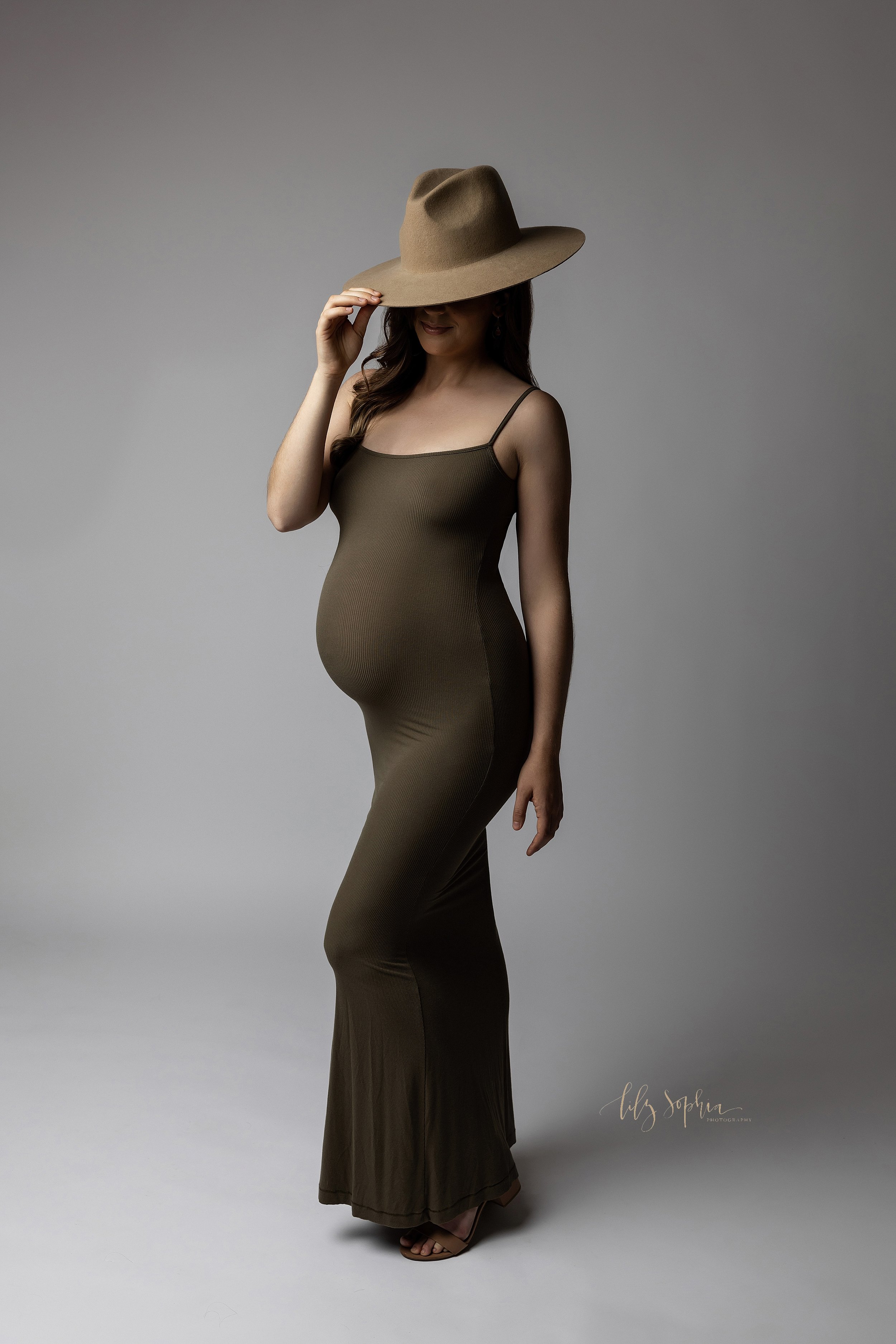  Modern maternity photo shoot with an expectant mother in a natural light studio with the mother holding onto the brim of a stetson hat and wearing a spaghetti strap full-length jersey gown taken near Midtown in Atlanta, Georgia. 