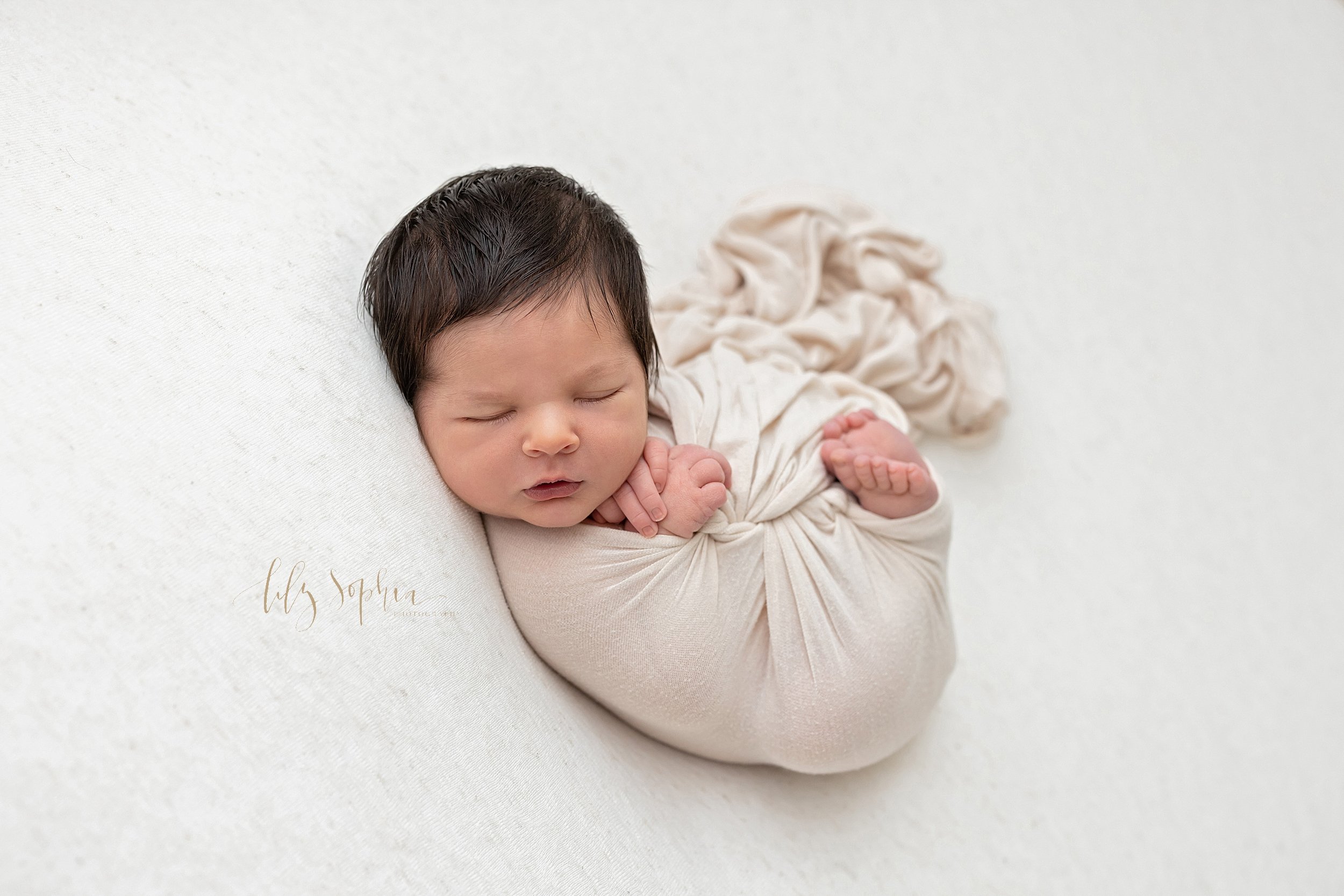  Newborn photo of a baby boy as he sleeps on his back wrapped as a bundle of joy in a stretchy swaddle with his hands and his feet peeking out taken near Ansley Park in Atlanta in a natural light studio. 