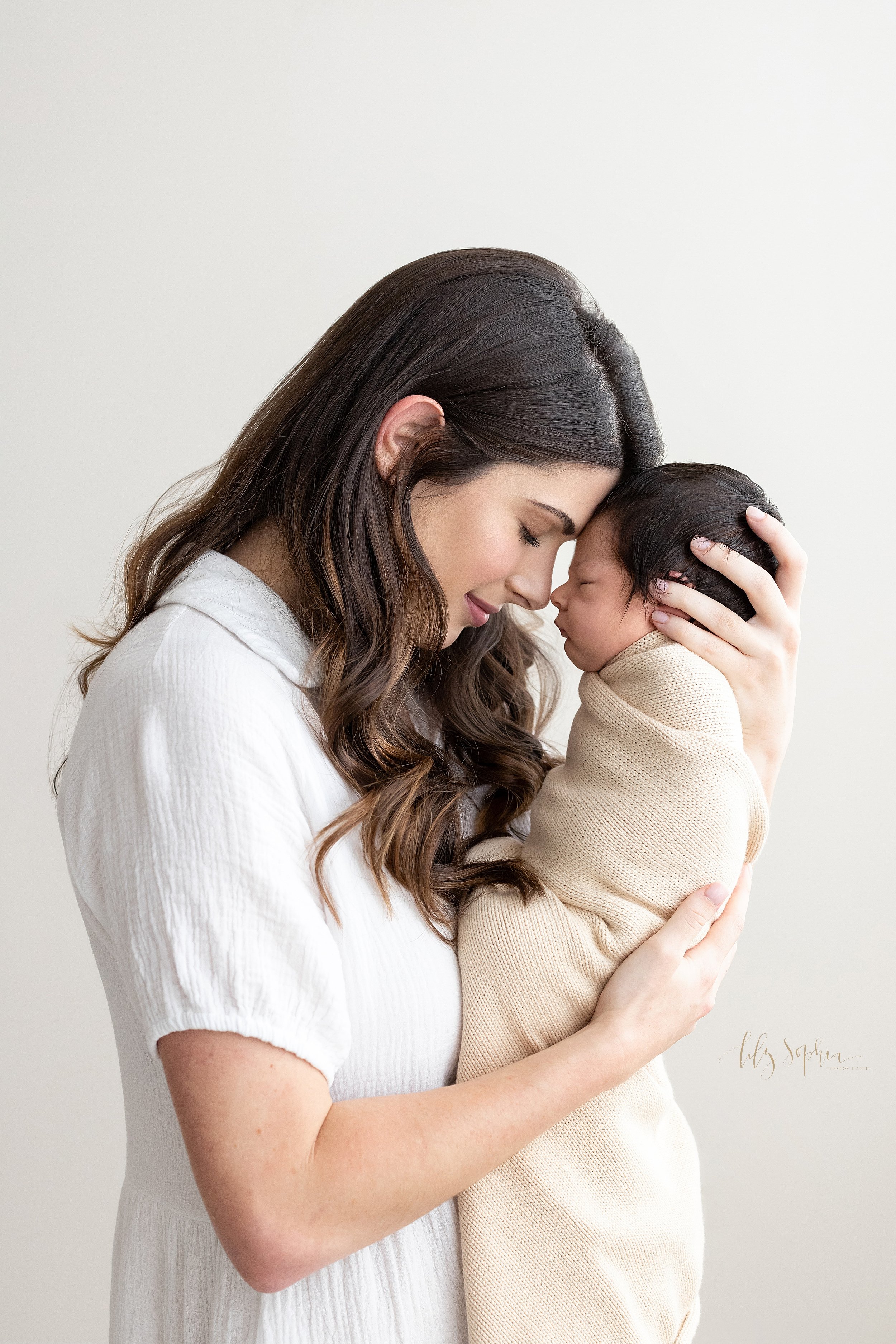  Newborn family photograph of a mother holding her sleeping infant son in front of her with her left hand holding his head and her right hand holding his bottom while she places her forehead against hers while standing in a natural light studio near 