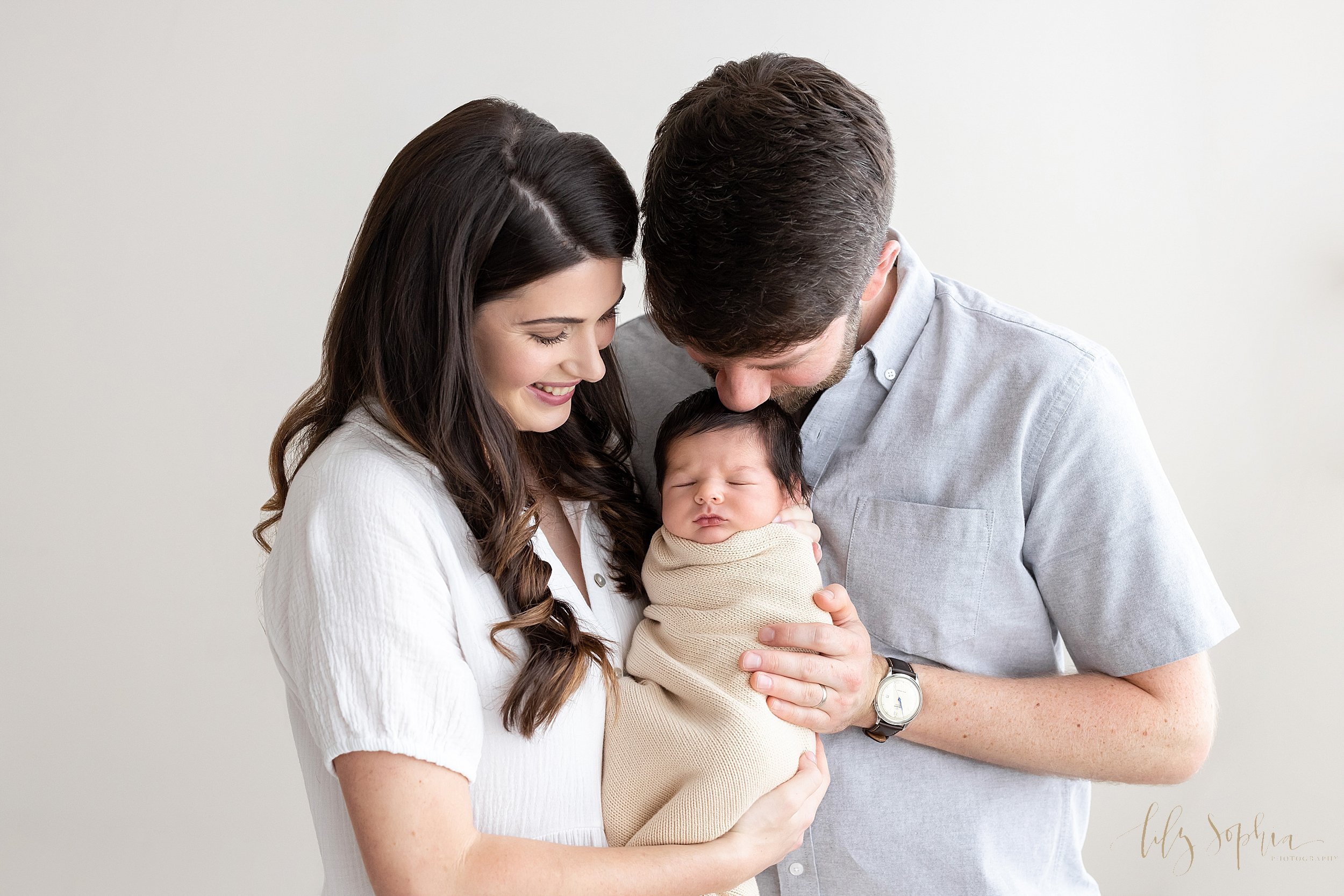  Newborn family pictures of a mother holding her sleeping newborn son swaddled to his chin in a natural blanket in her arms in front of her husband’s chest as he kisses the top of his infant son’s head taken in natural light in a studio near Buckhead