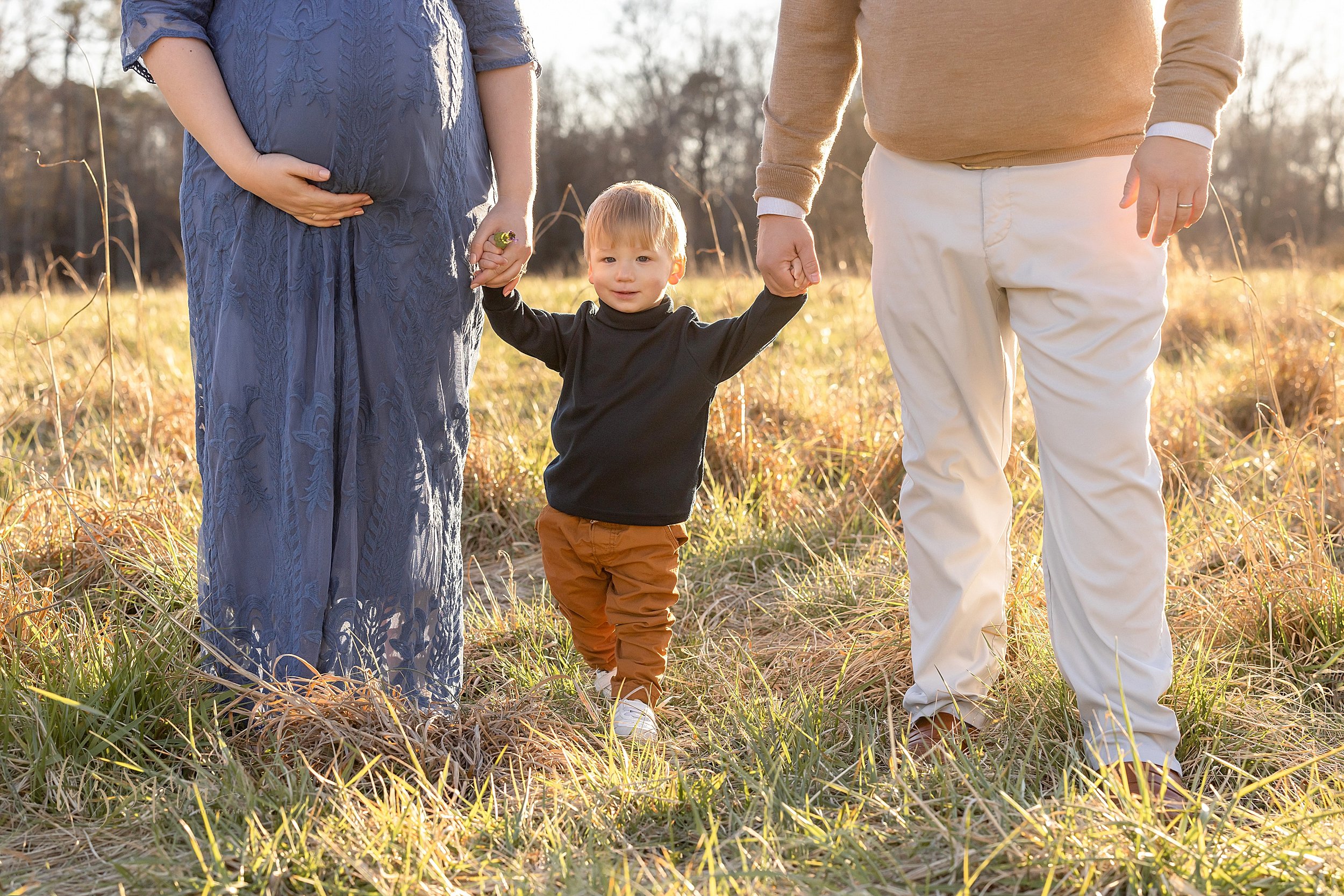  Fall family maternity pictures of a pregnant mother and her husband walking with their young son between them as the couple hold his hands and mom holds the base of her belly with her right hand while they walk through a field near Atlanta at sunset