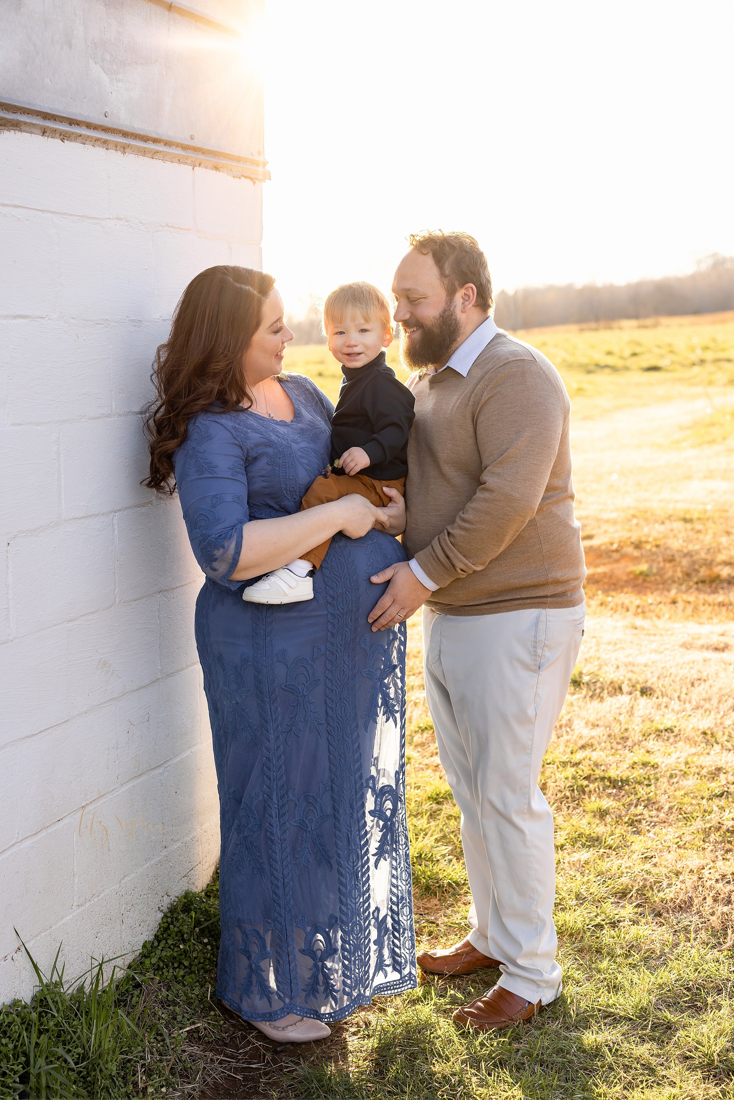  Fall family maternity photos of a pregnant mother holding her young son as her husband faces her with his right hand on her belly taken at sunset near a field in Atlanta. 