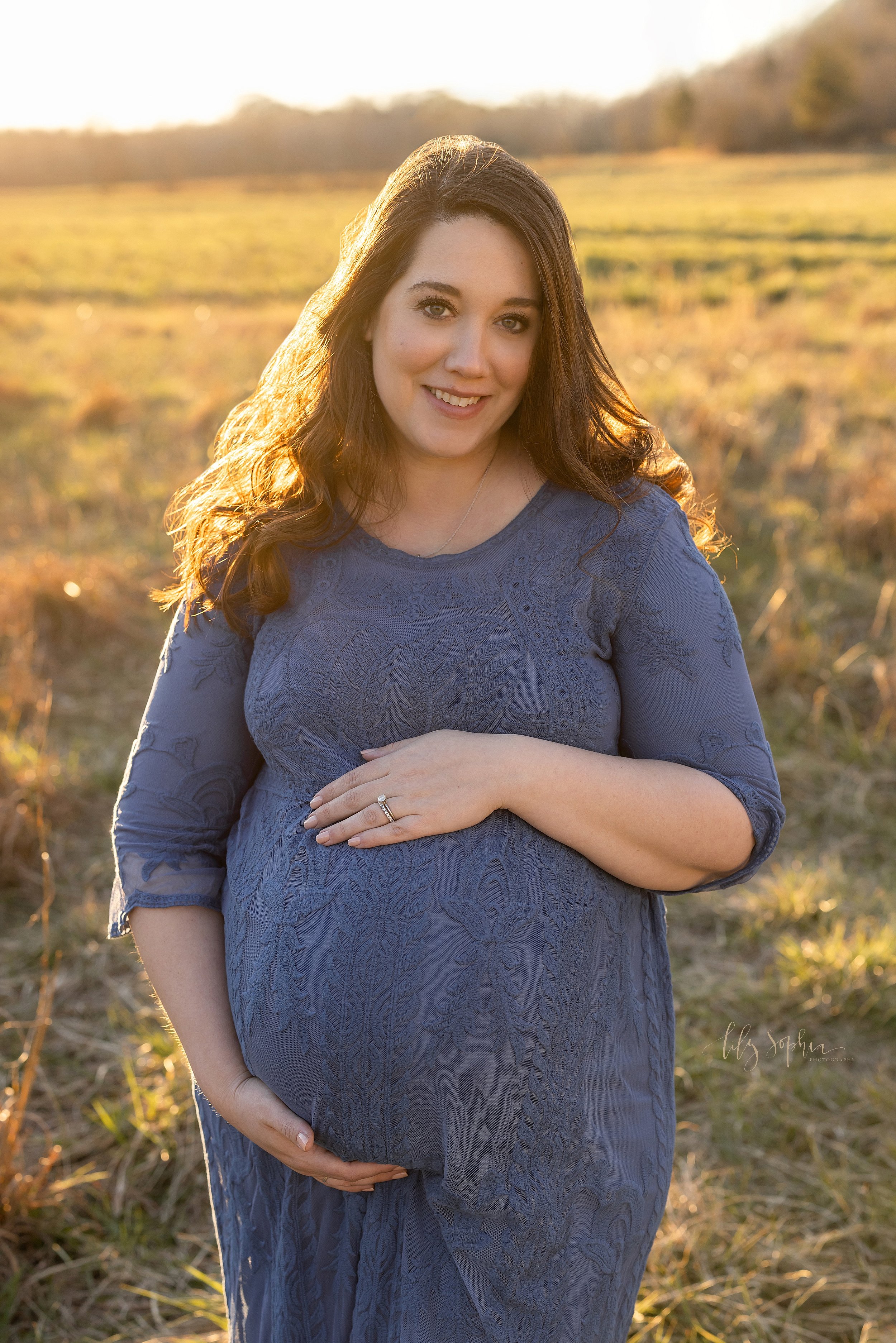  Fall maternity photoshoot of an expectant mother as she stands in a long sleeved blue scoop-necked dress and frames her belly with her hands at sunset during autumn in a field near Atlanta. 
