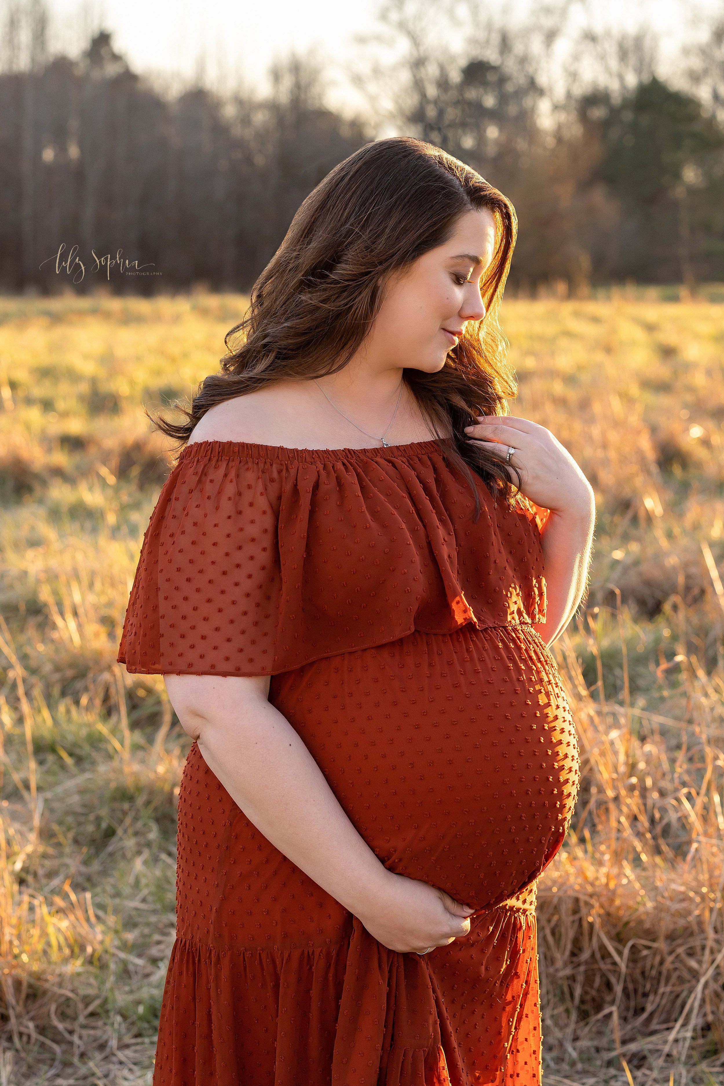  Fall maternity picture of a pregnant mother as she stands wearing a rust swiss dot off the shoulder wide ruffled bodice full-length cotton gown as she strokes her hair with her left hand and holds the base of her belly with her right hand taken in a