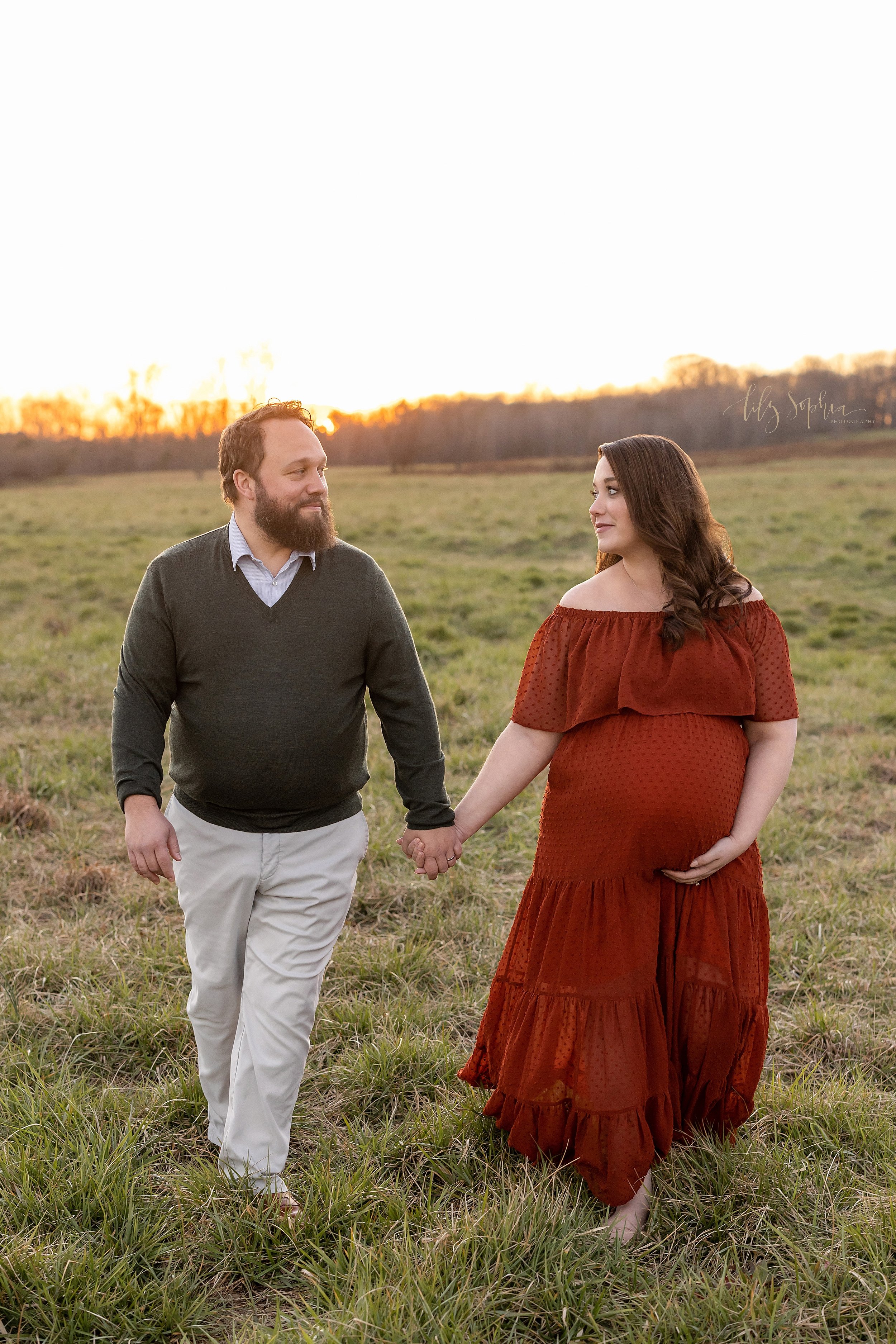  Fall maternity photoshoot of an expectant couple as the husband holds his wife’s right hand while walking through a field near at sunset near Atlanta, Georgia. 