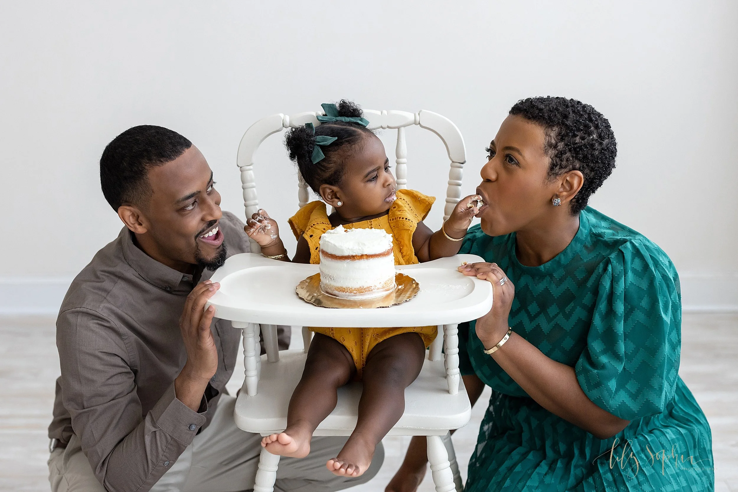  Family first birthday picture of a dad squatting on the left side of his daughter sitting in an antique high chair with her smash cake on the tray and mom sitting on the right side of her daughter as the one year old puts icing into her mom’s mouth 