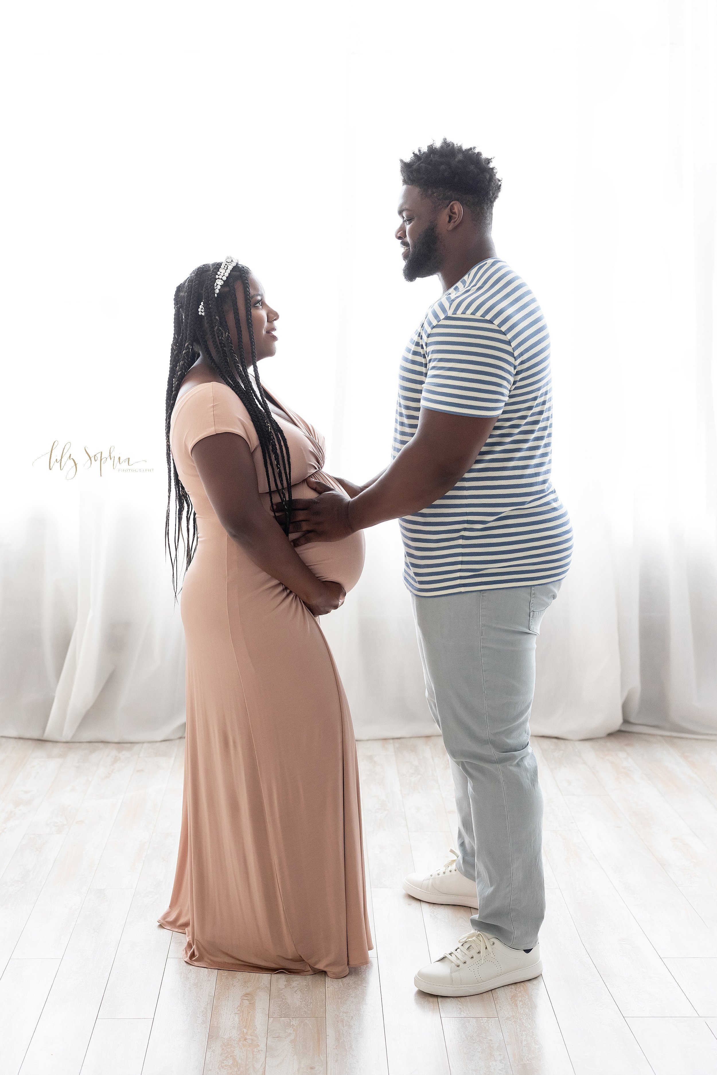 Maternity picture of an African-American expectant couple as they stand in front of a window streaming natural light and face one another with the husband placing his hands on his child in utero while the wife holds the base of her belly taken near 