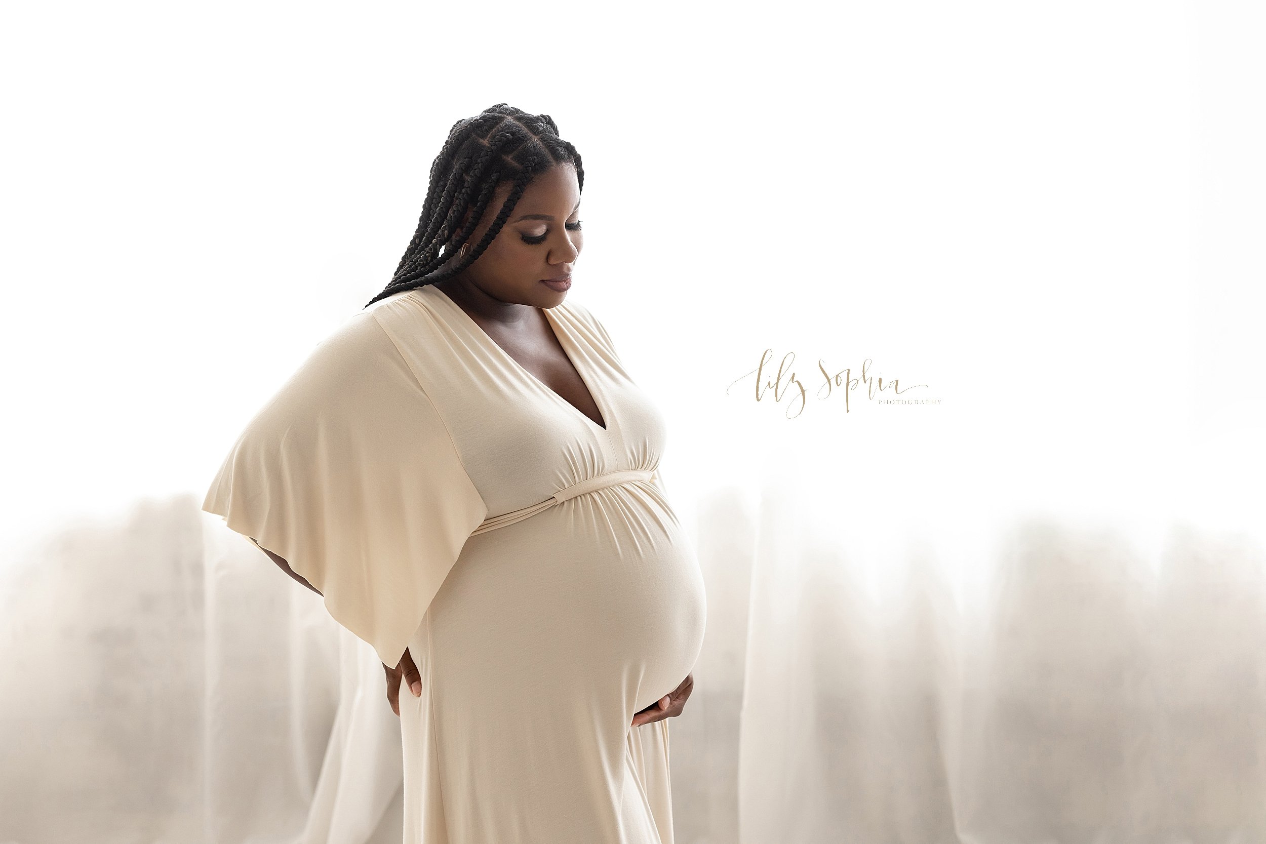  Maternity photos of a black pregnant woman as she stands with her left hand beneath her belly and her right hand on the back of her hip taken in a studio in front of a window streaming natural light near Kirkwood in Atlanta, Georgia. 