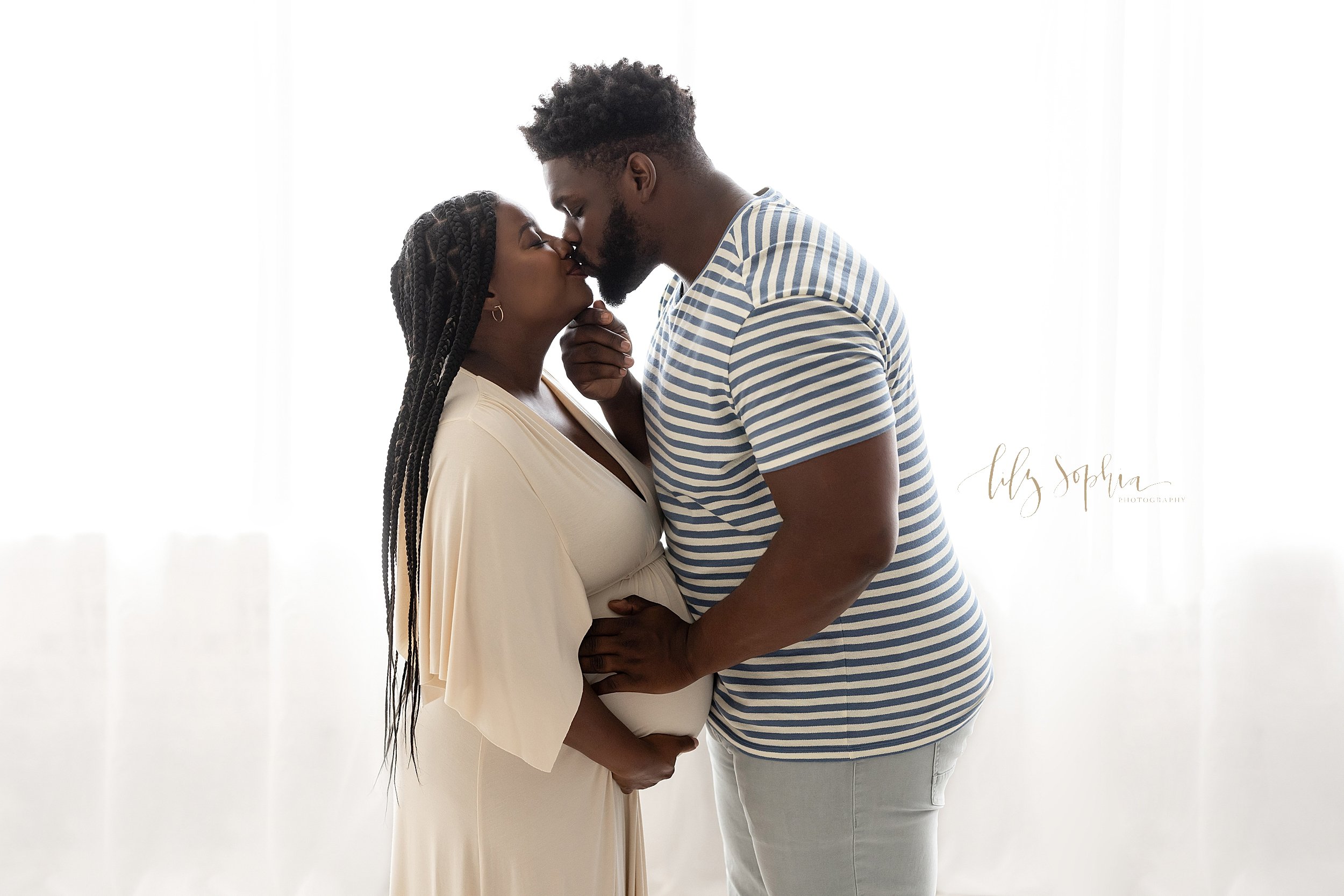  Maternity photoshoot of a black expectant couple with them facing one another in front of a window streaming natural light as the husband places his right hand under his wife’s chin and kisses her taken in a studio near Morningside in Atlanta. 