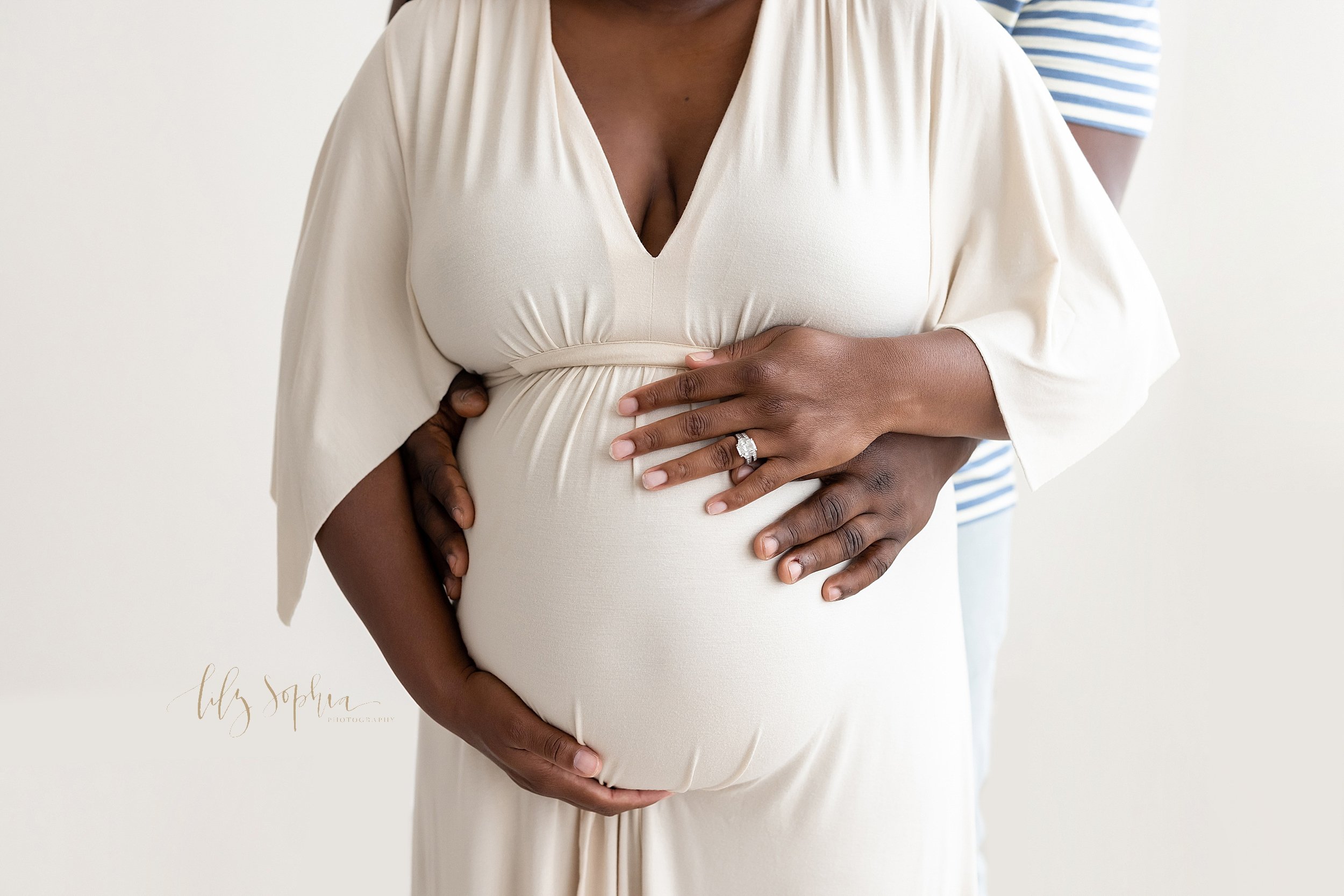  Close-up maternity picture of black expectant parents with the father standing behind his wife and reaching to hold his child in utero as the mother frames her belly with her hands taken in a natural light studio near Midtown in Atlanta. 