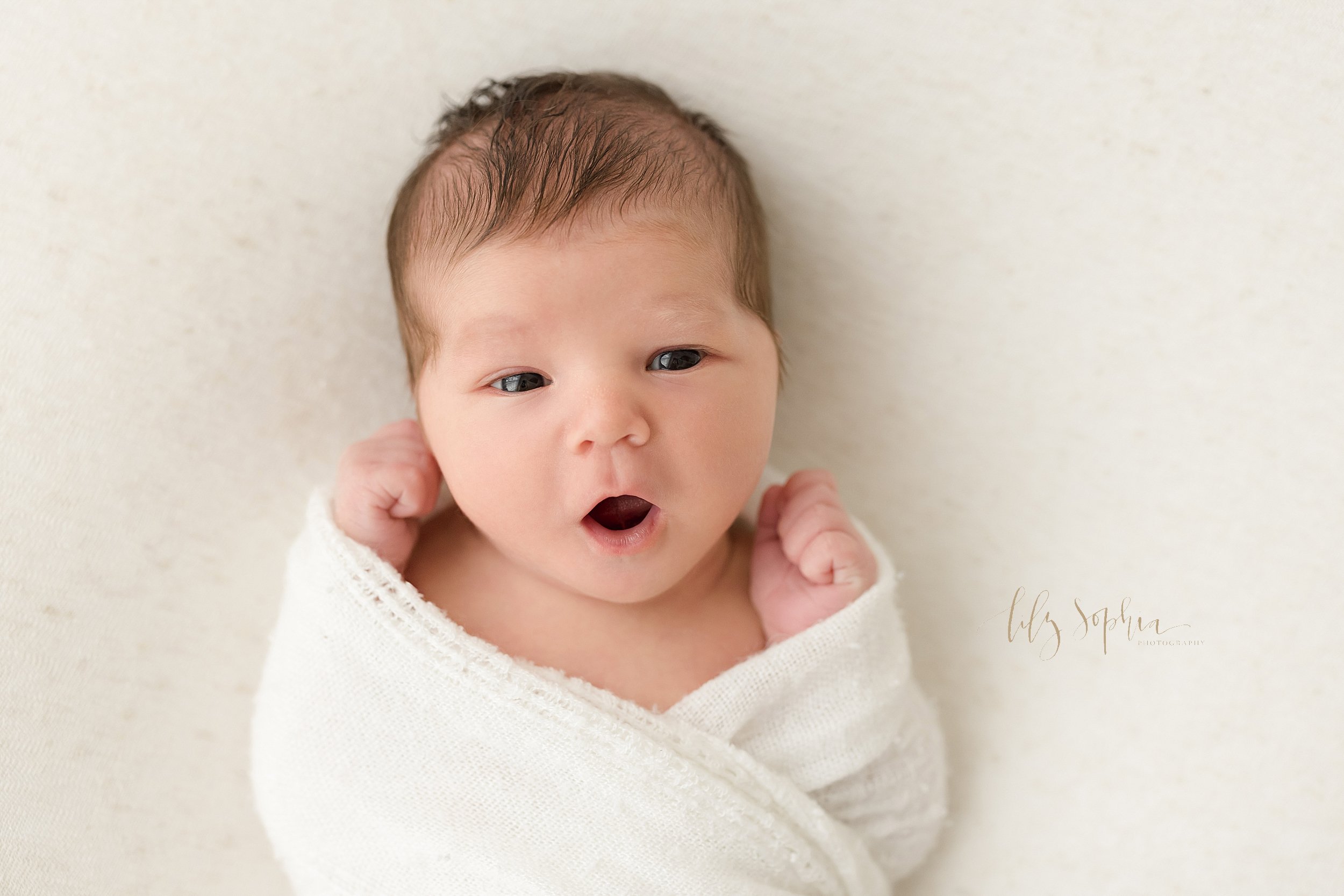  Newborn picture of an infant boy lying on his back with a white blanket wrapped around him as he opens his mouth with his eyes opened wide taken in a studio near Poncey Highlands in Atlanta that uses natural light. 