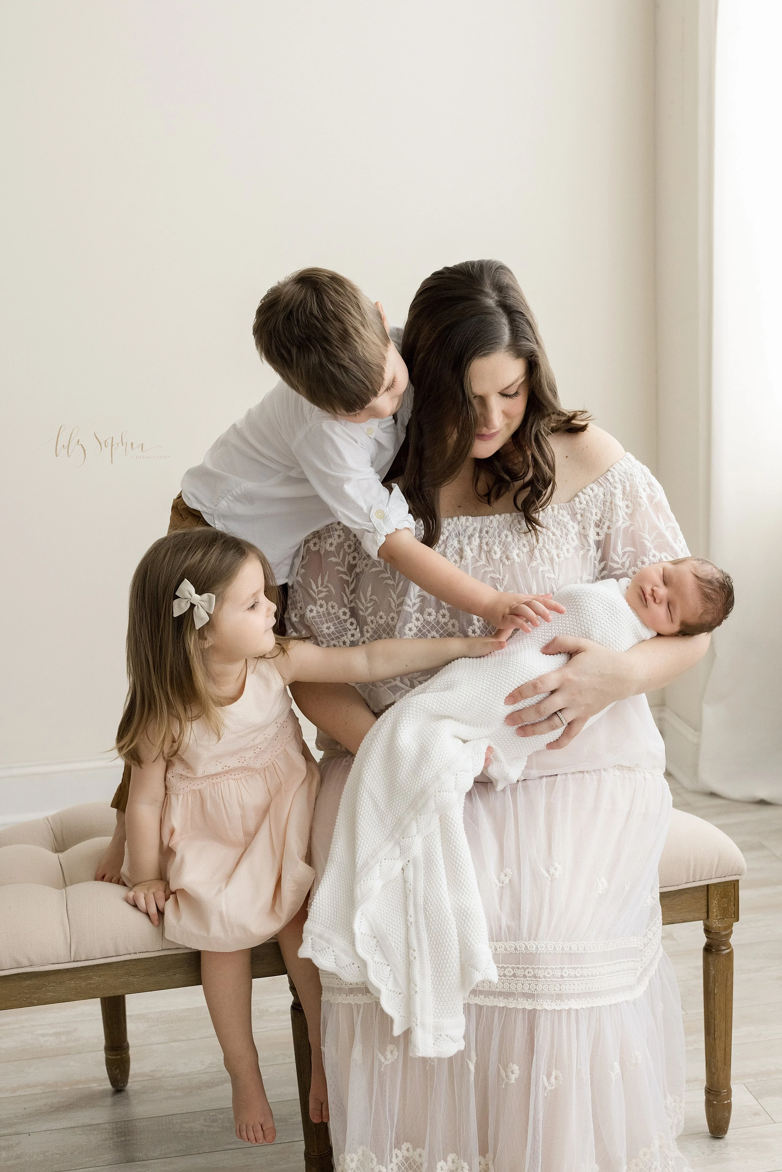  Family newborn picture of a mother sitting on a tufted bench holding her infant son in her arms as her young daughter sits next to her and places her hand on her brother and her young son stands on the bench next to her and reaches over her shoulder