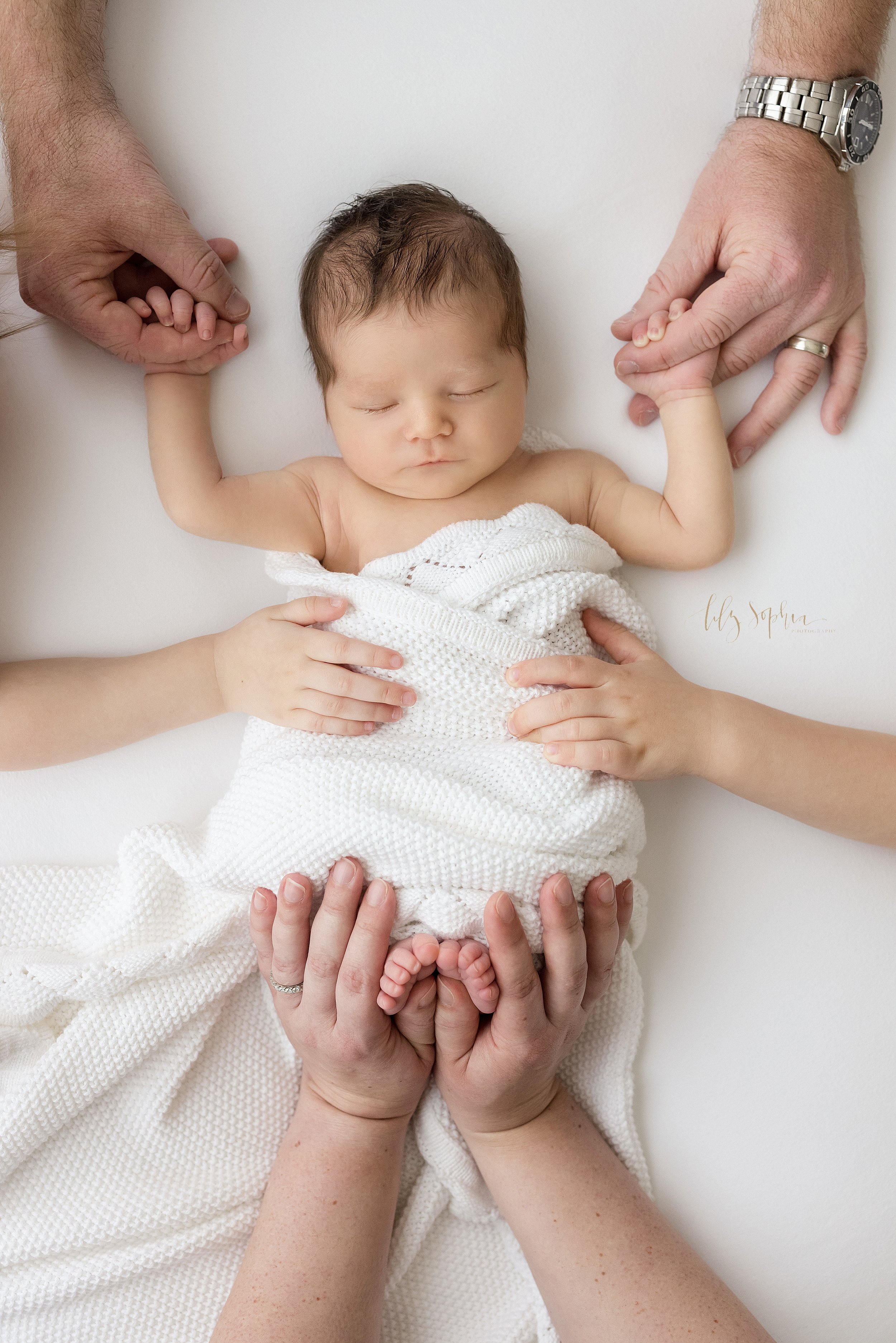  Family newborn photo of a sleeping newborn baby boy with his hands above his head to hold his father’s index fingers, his tiny feet sticking out of the soft white crocheted blanket snuggling him as his mom hold his feet and his siblings lay their ha