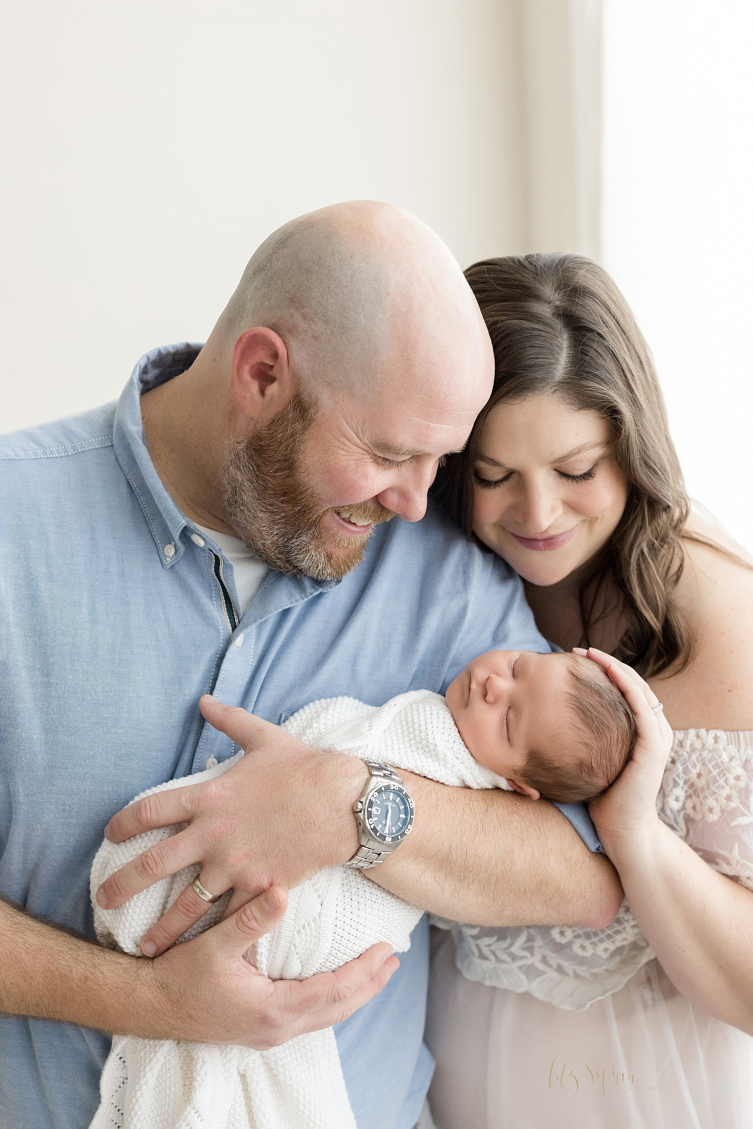  Newborn photoshoot with dad holding his newborn baby boy in his arms as mom stands slightly behind dad on his right hand side and she places her right hand on her son’s head as the couple stand in front of a window streaming natural light to admire 