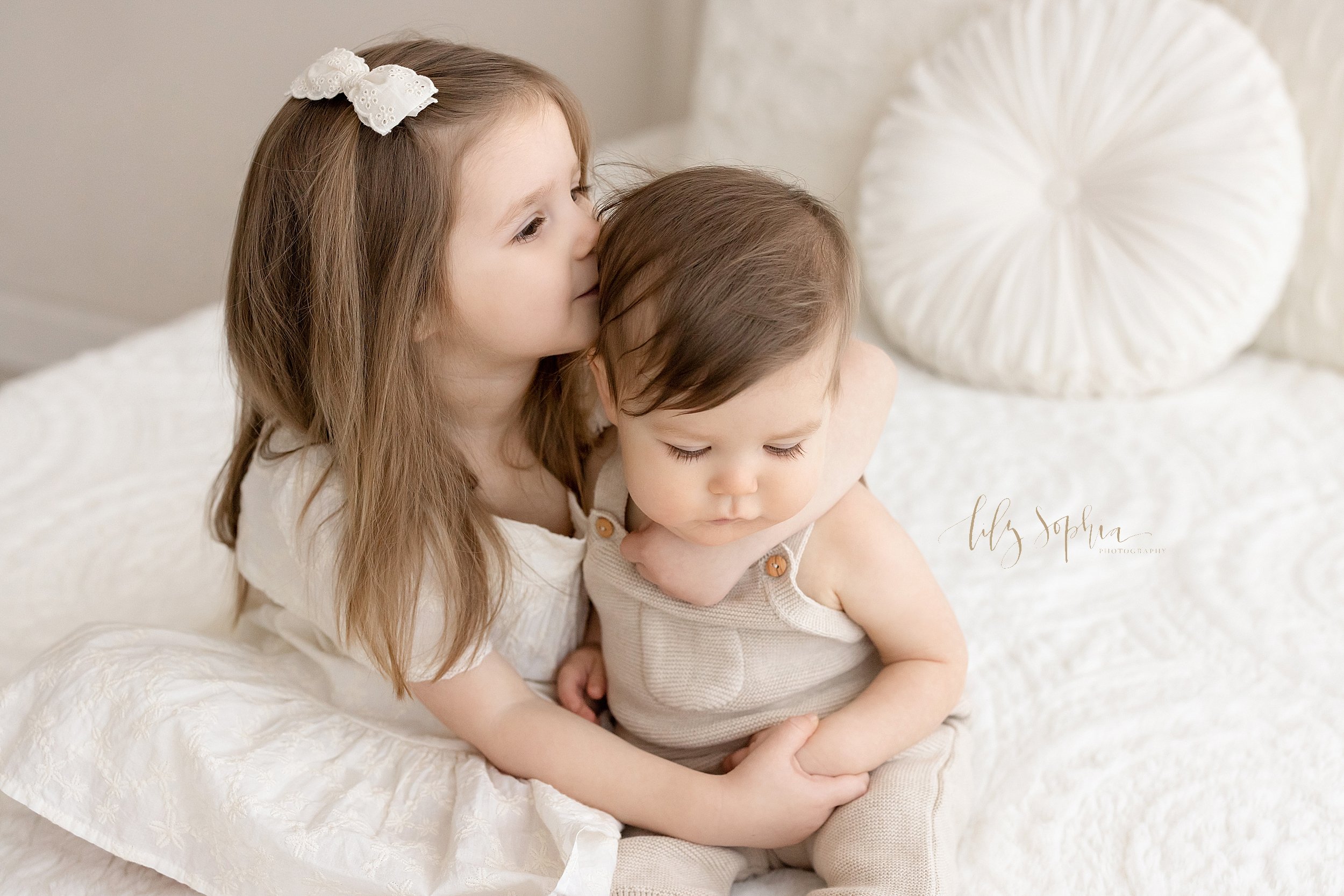  Sibling photo of a big sister sitting next to her one year old brother atop a bed as she holds his hand and kisses him on the side of his head taken in a natural light studio near Buckhead in Atlanta, Georgia. 