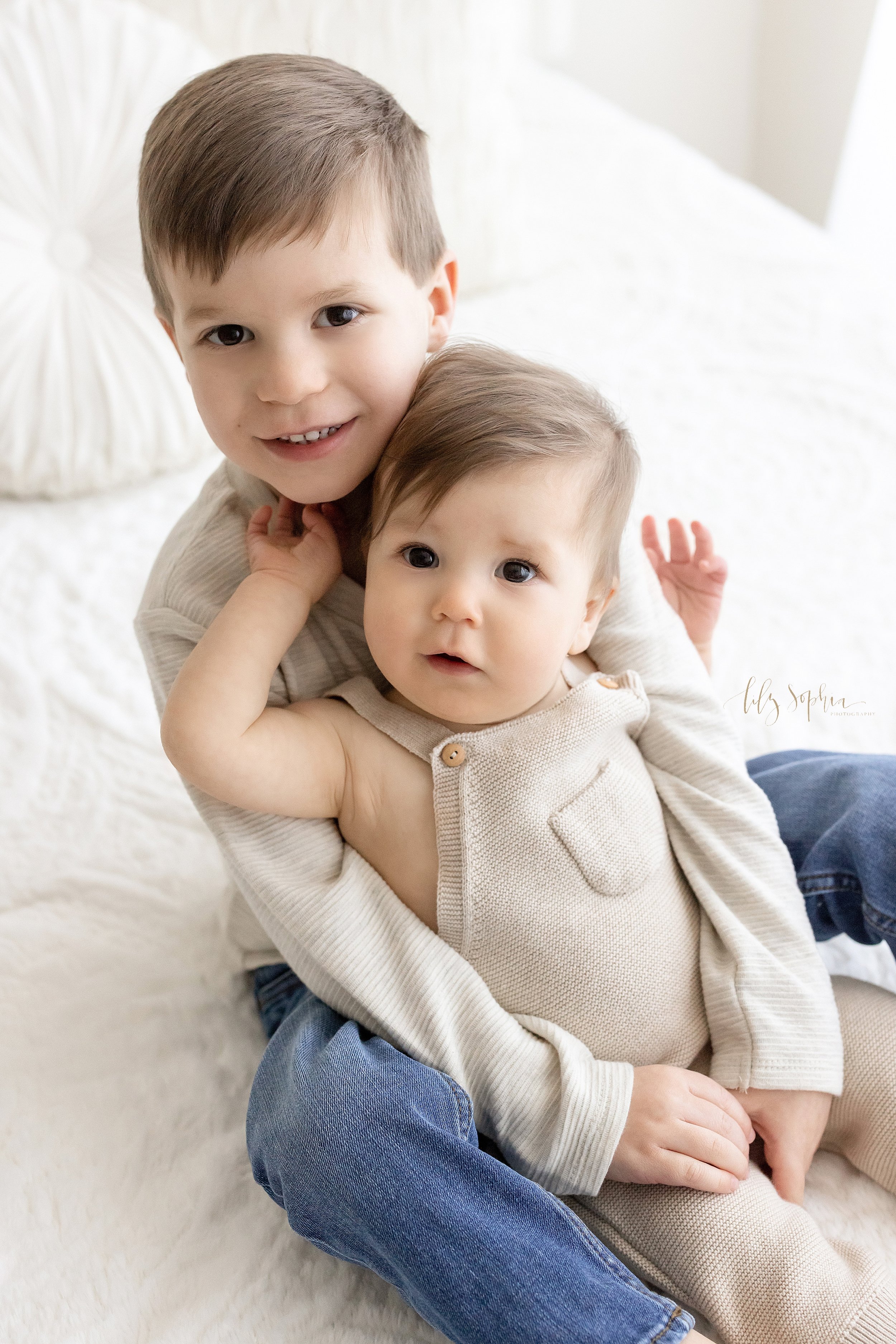  Sibling photo of two brothers sitting on a bed the older brother has his one year old brother sitting between his legs as he wraps his arms around him taken near Virginia Highlands in Atlanta in a natural light studio. 