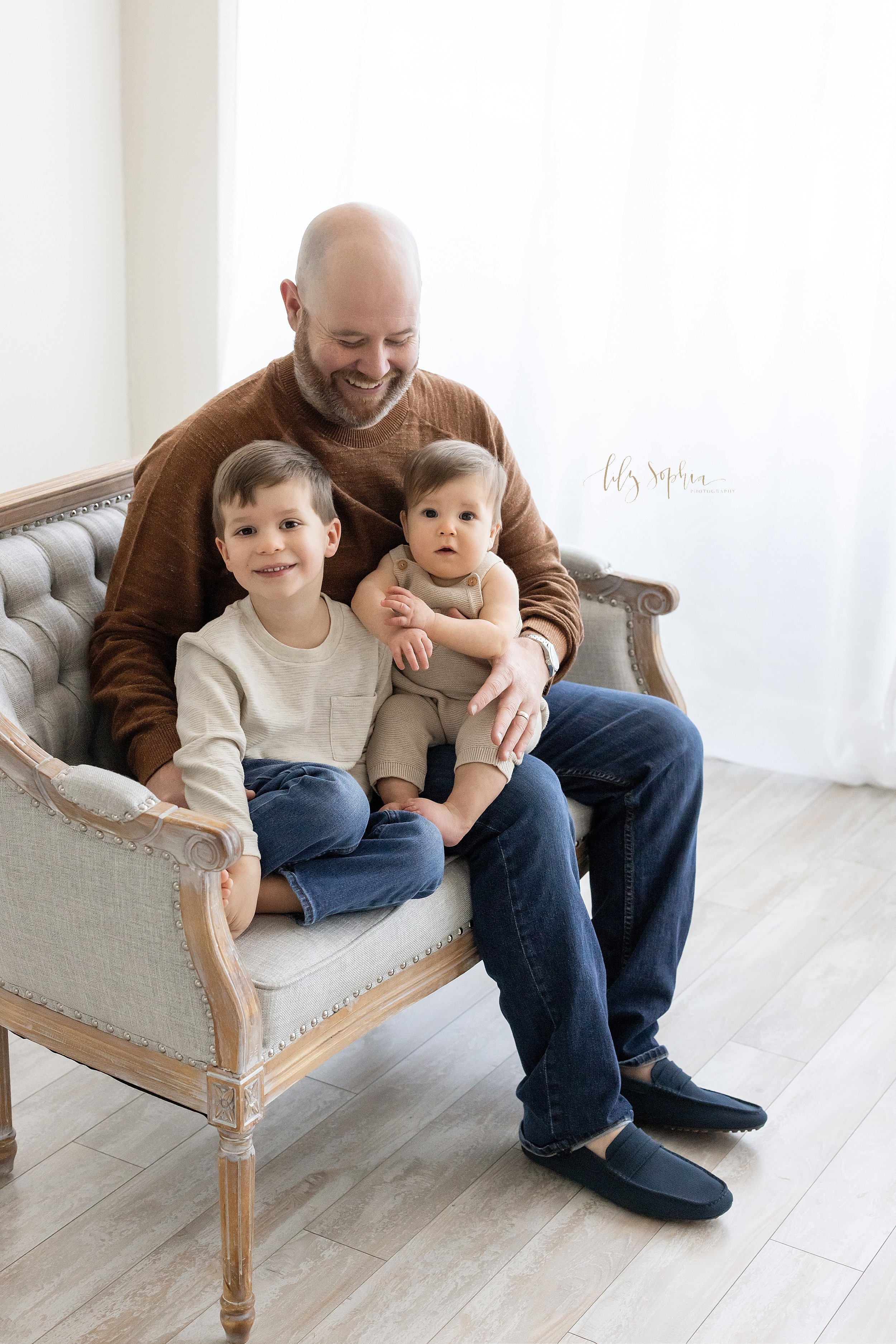  Family photo of a father with his one year old son on his lap as his older son sits next to him on a tufted nail trimmed sofa in front of window streaming natural light in a studio near Kirkwood in Atlanta. 