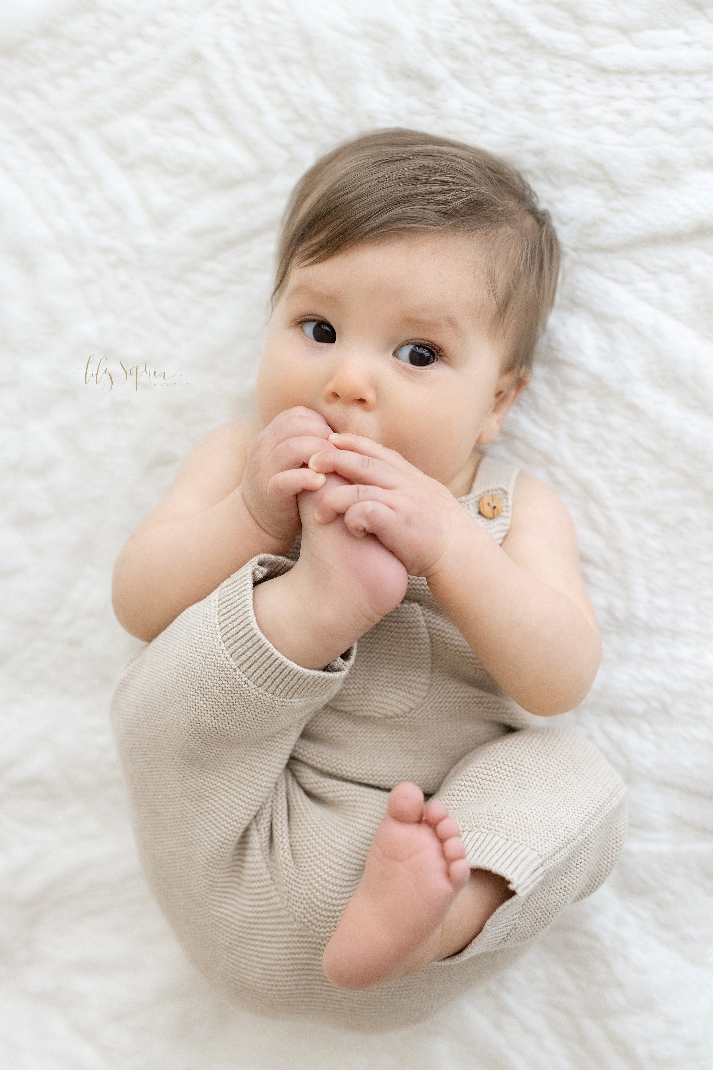  First year photo of a baby boy lying on his back in a natural light studio chewing on his toes taken near Midtown in Atlanta, Georgia. 