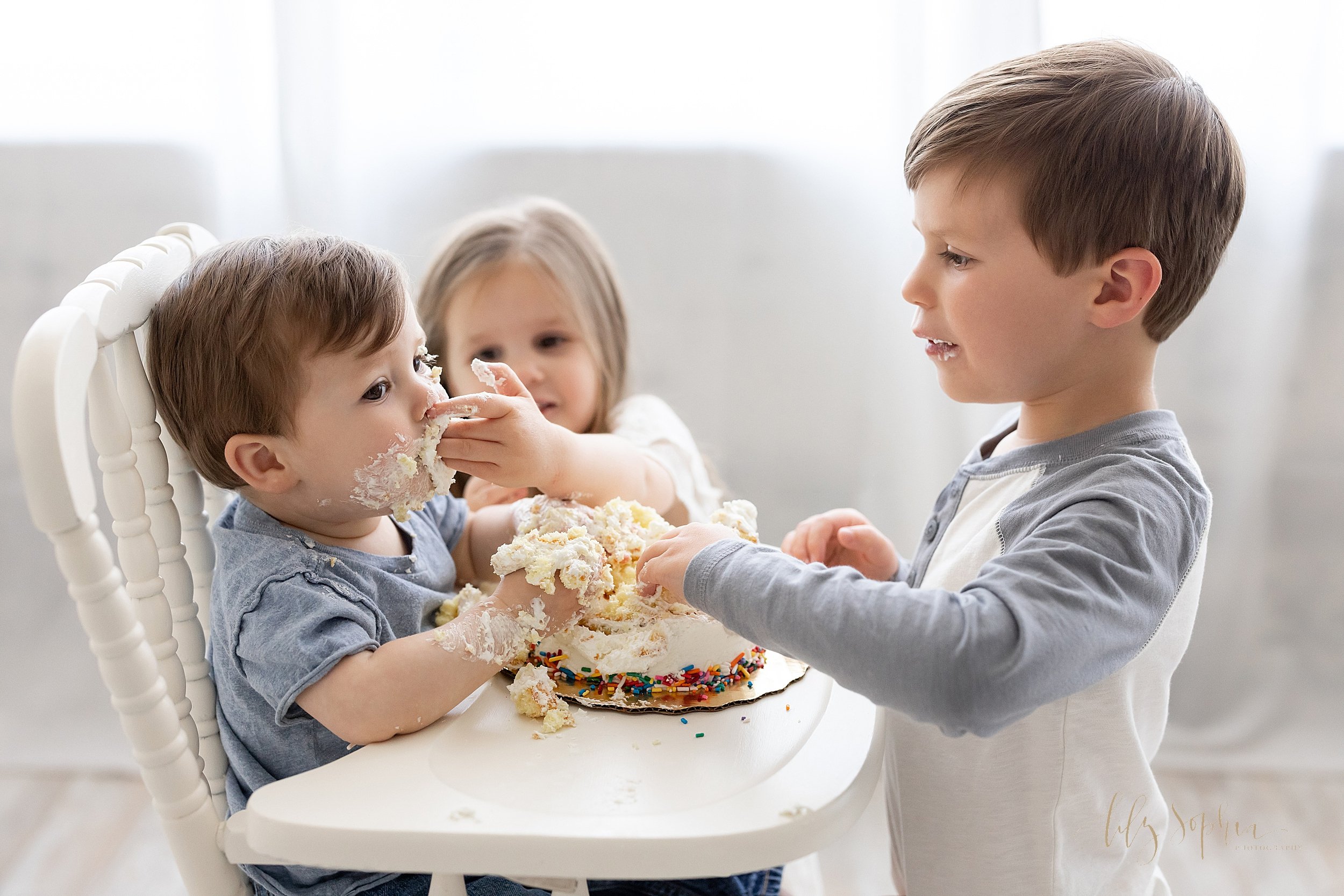  First birthday family smash cake photo of a one year old boy sitting in an antique high chair as his sister places icing in his mouth and his brother stands in front of him to get another bite to give him taken in front of a window streaming natural