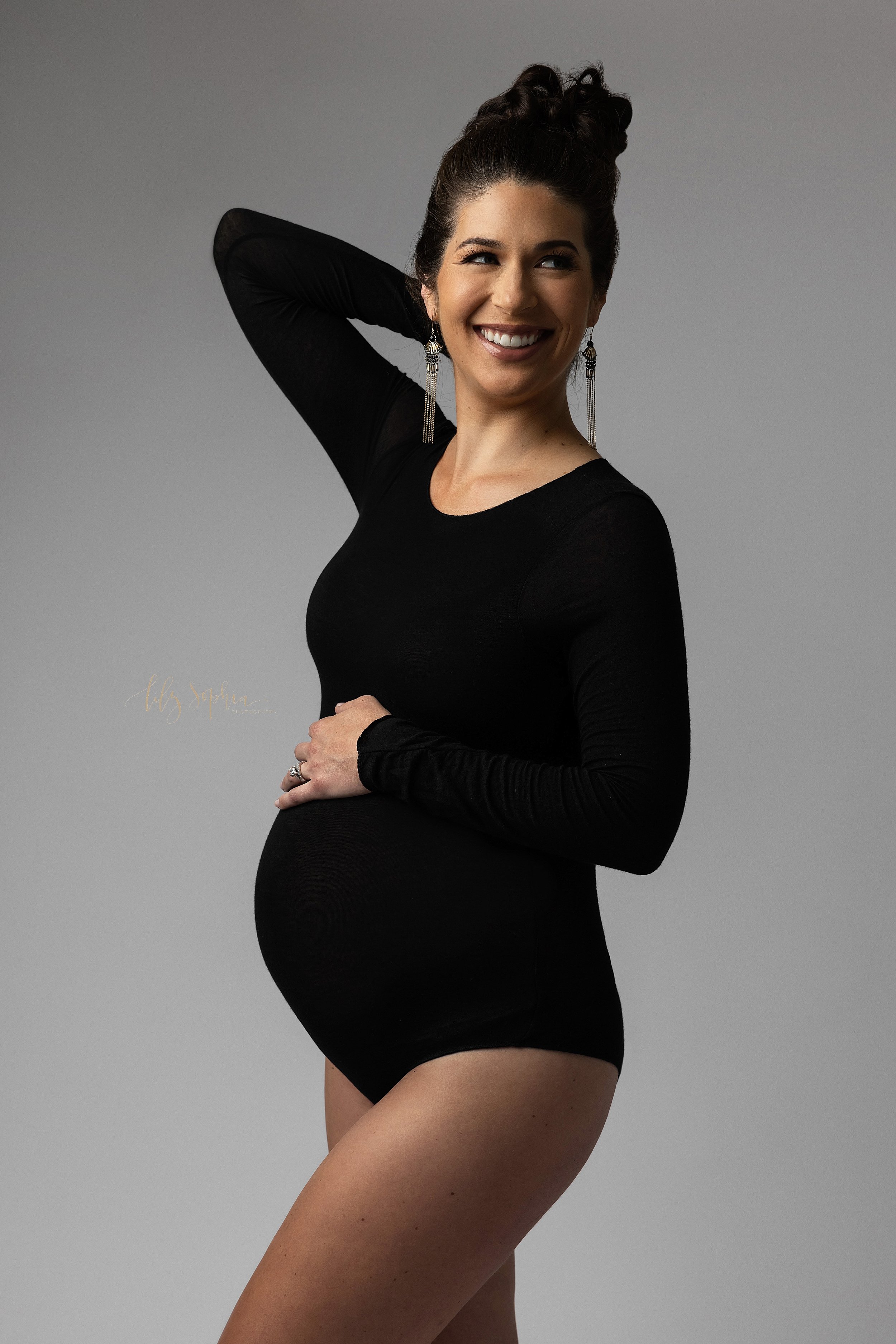  Modern maternity photoshoot of an expectant mother as she celebrates being pregnant while wearing a black scoop necked long sleeved body suit and places her left hand on top of her belly while placing her right hand behind her head as she turns to l