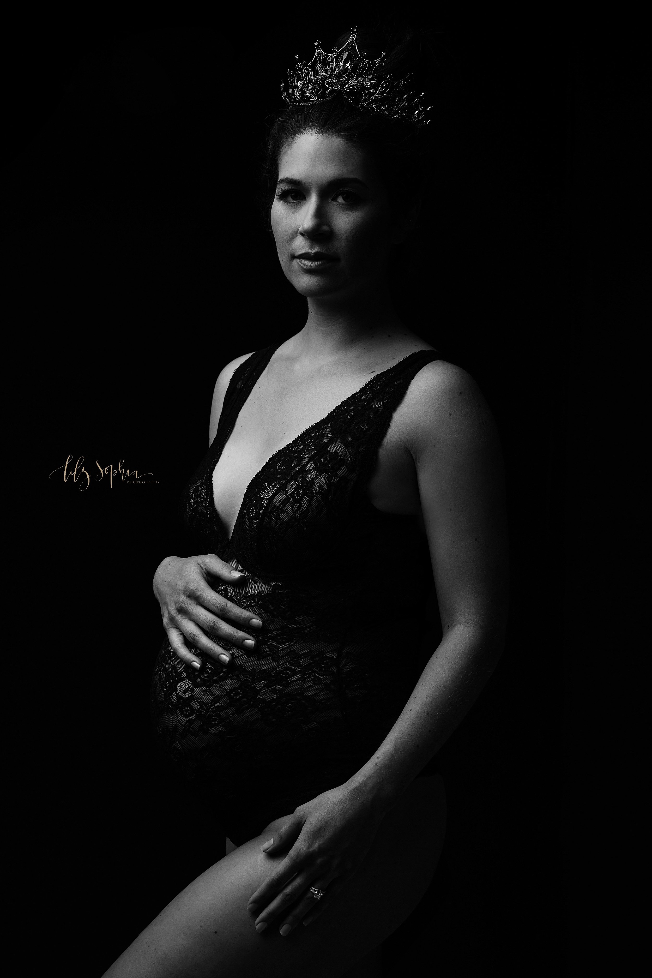  Modern maternity photoshoot with an expectant mother wearing a crown on her head and a lace plunging necked bodysuit as she stands with her right hand on the top of her belly and her left hand on her left thigh taken near Sandy Springs in Atlanta in