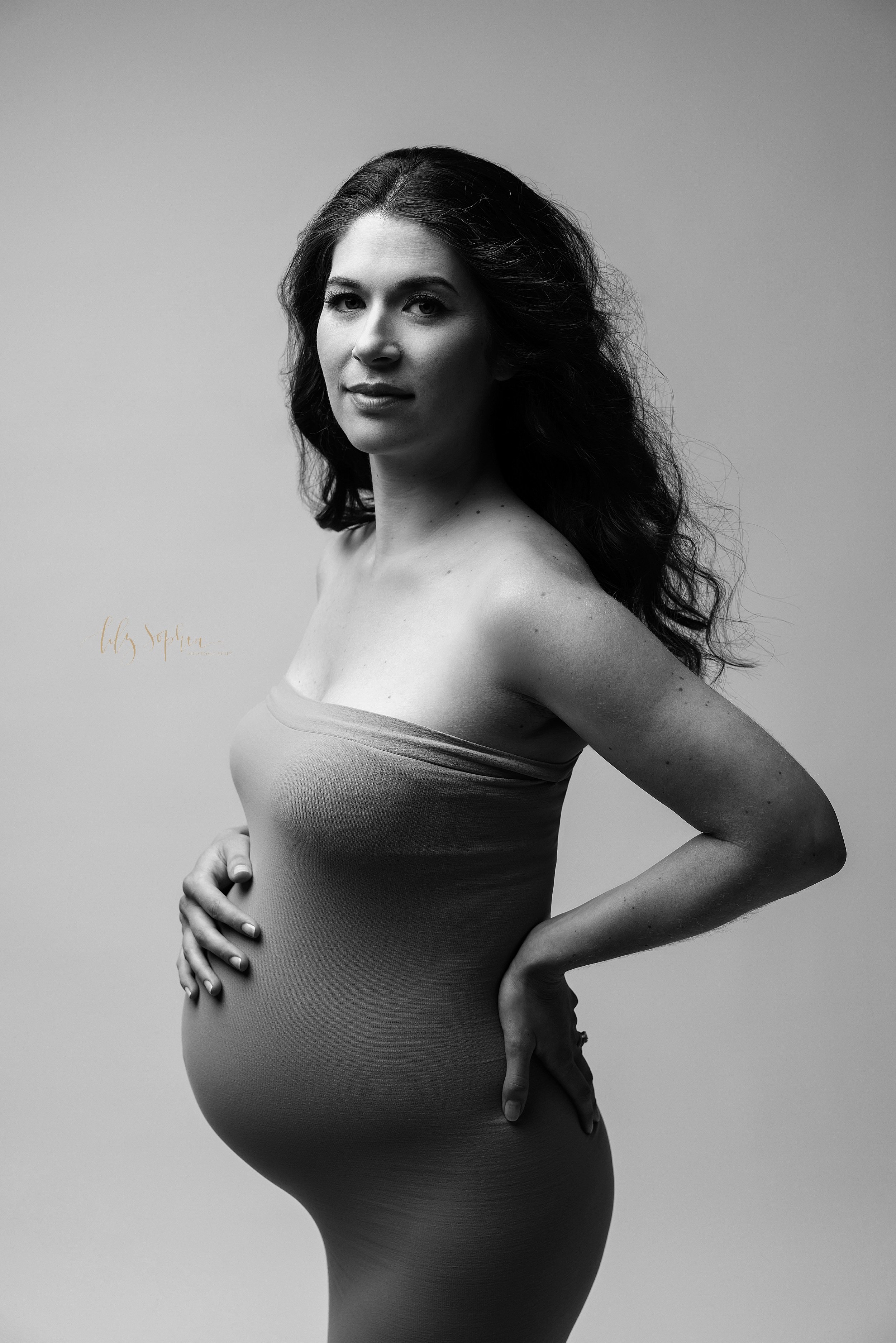  Modern maternity picture of an expectant woman as she stands with her right hand at the top of her belly and her left hand on the back of her hip taken near Kirkwood in Atlanta in a photography studio. 