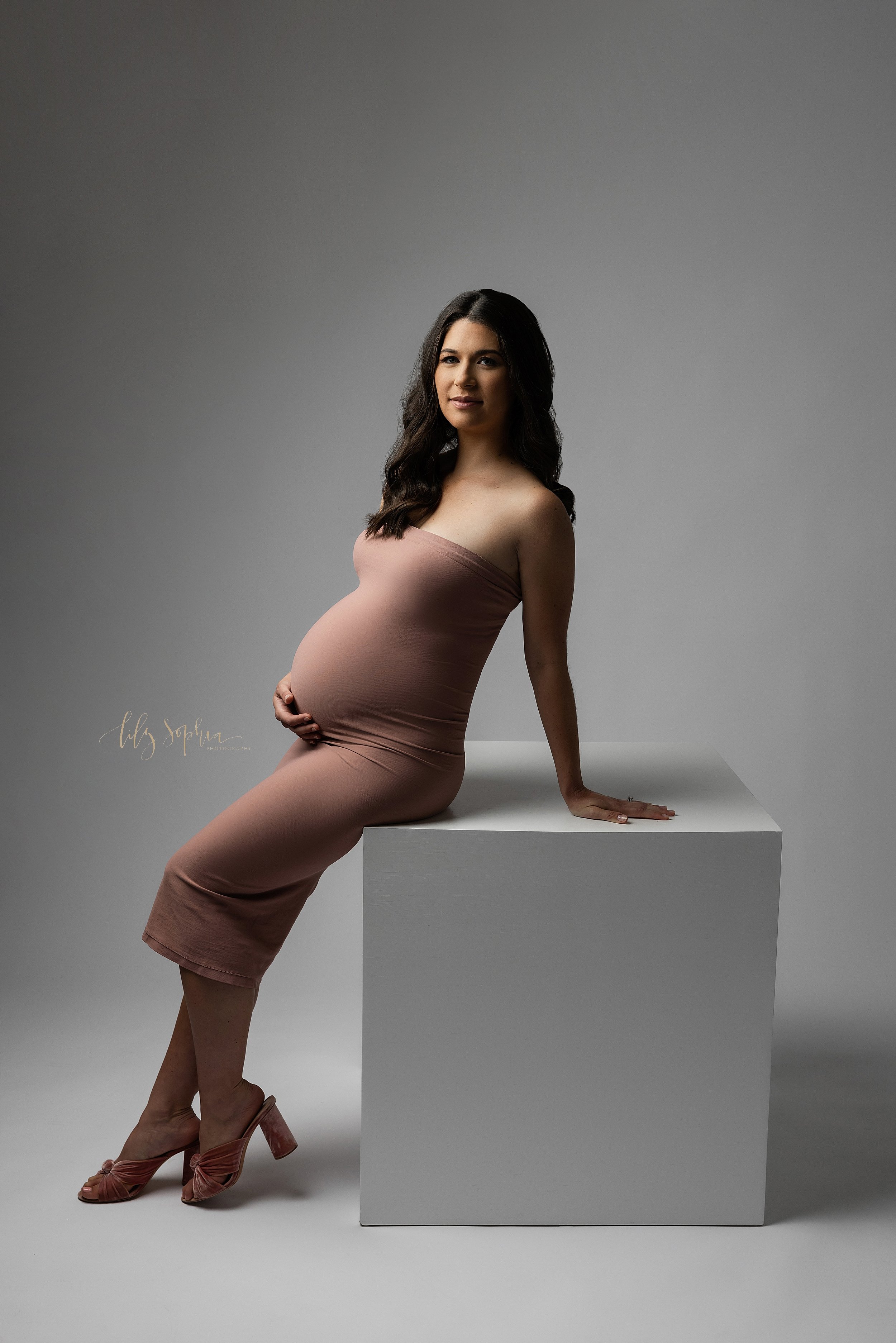  Modern maternity photoshoot of a pregnant woman wearing a strapless jersey knit gown as she leans against a white cube and rests on her right arm taken in a studio near Midtown in Atlanta. 