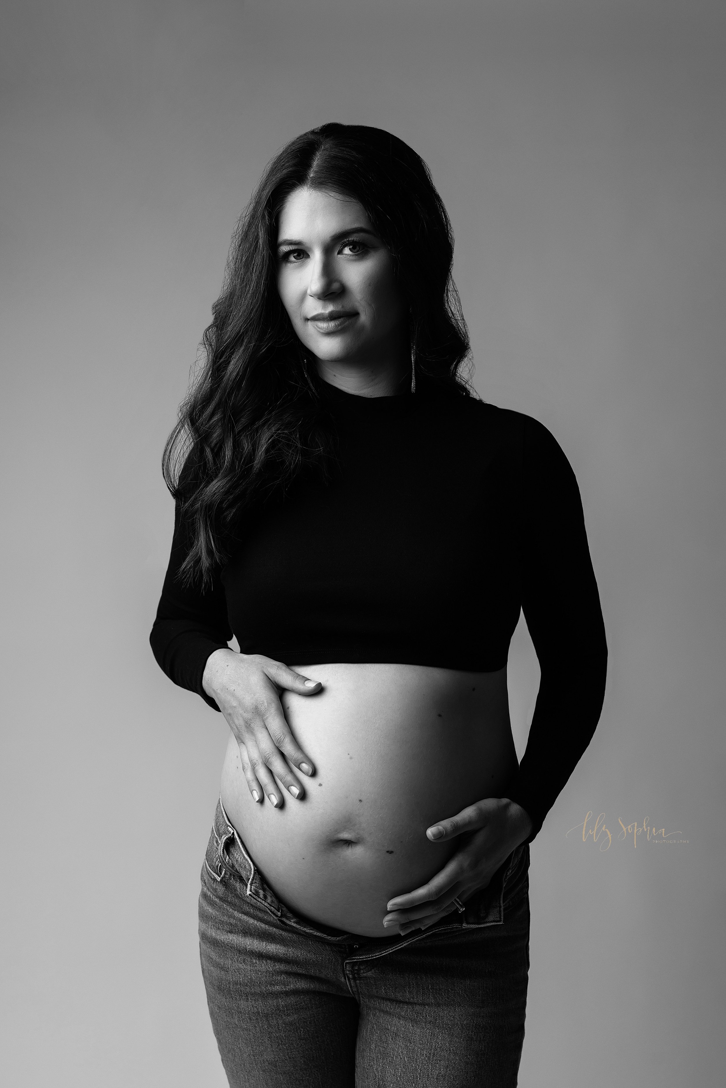  Modern maternity photoshoot of a woman wearing a cropped long sleeved shirt and unzipped blue jeans to bare her belly as she places her hands on her child in utero taken in a photography studio in Ponce City Market in Atlanta, Georgia. 