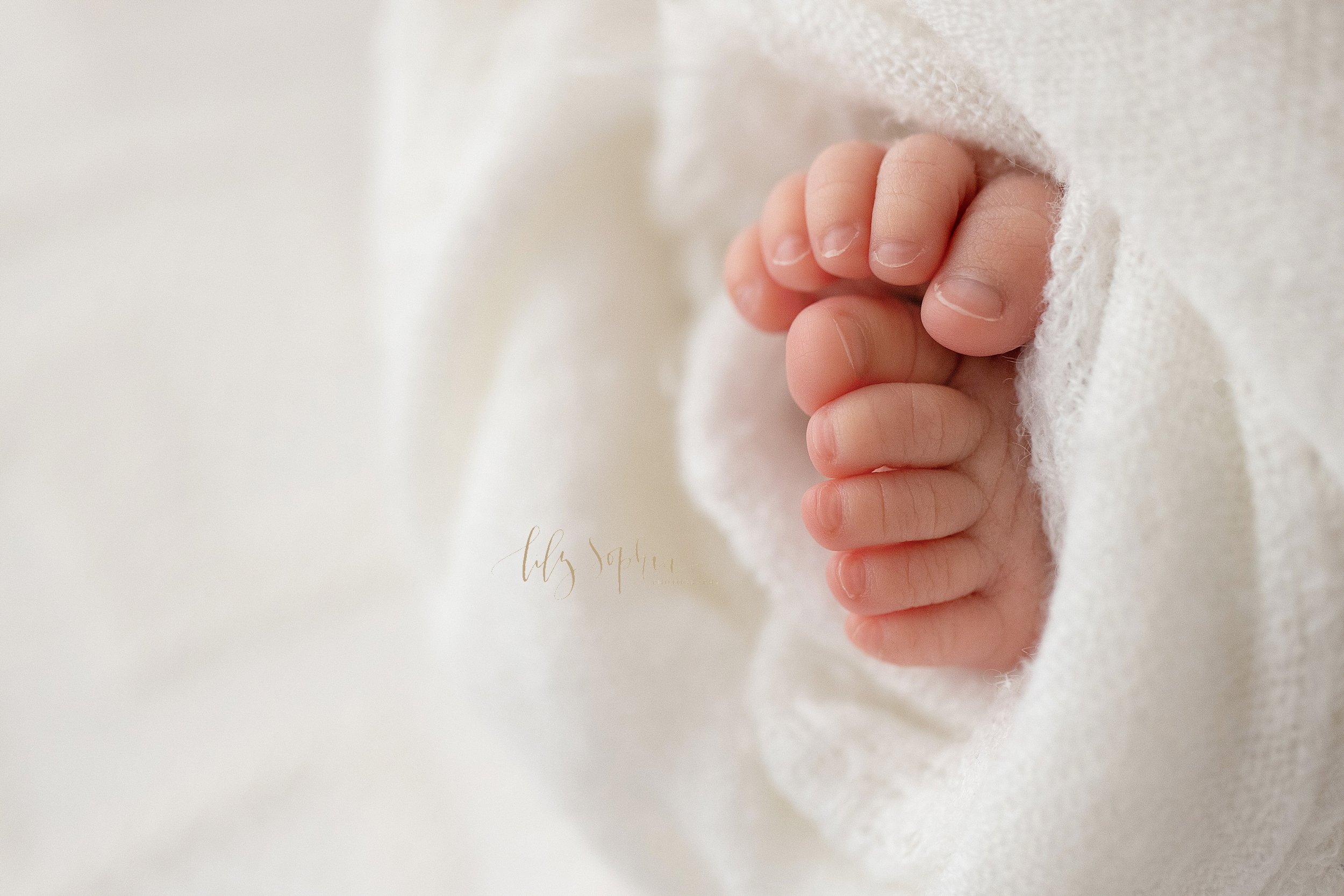  Newborn photoshoot of a newborn baby boy’s tiny toes as they peek out from a soft white blanket taken in a natural light studio near Cummings in Atlanta, Georgia. 