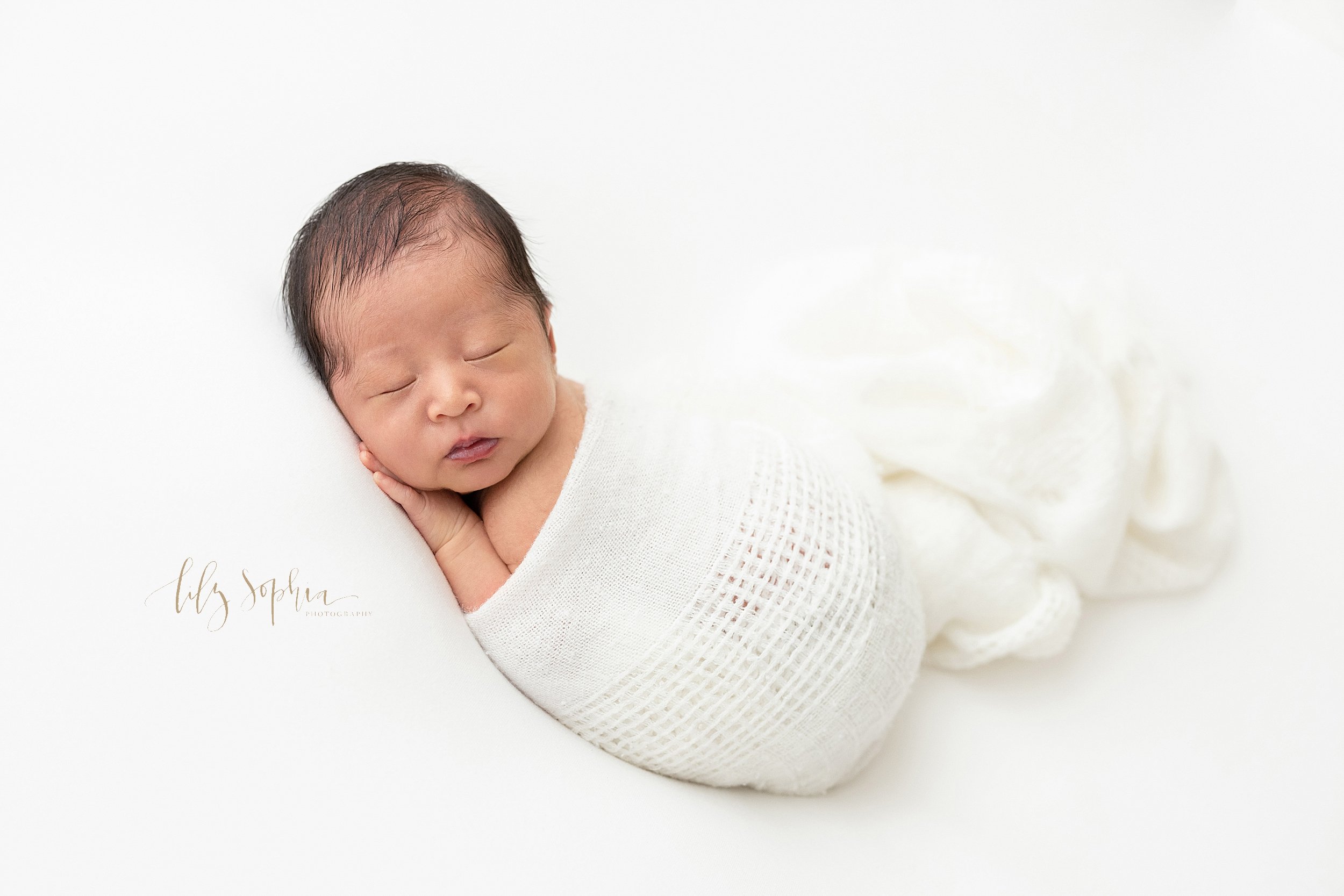  Newborn photoshoot of an Asian baby boy as he sleeps on his stomach with his head resting on his left hand as he looks over his left shoulder with a soft white blanket around him and flowing next to him taken near Morningside in a natural light stud