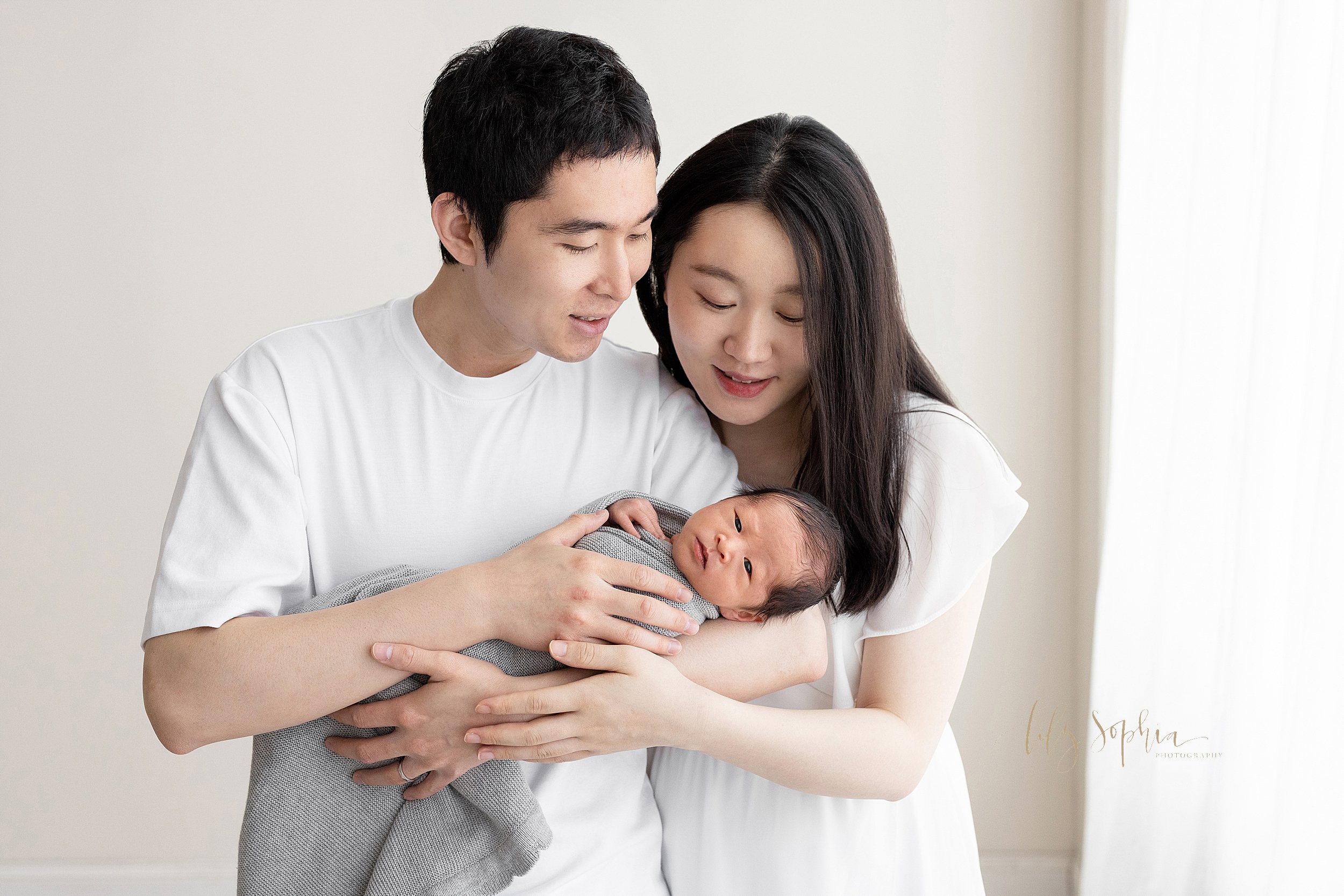  Newborn family photo of an Asian father holding his awake son in his arms as his wife stands slightly behind her husband’s left shoulder and peers over it at her newborn son as the couple stand in front of a window streaming natural light in a studi
