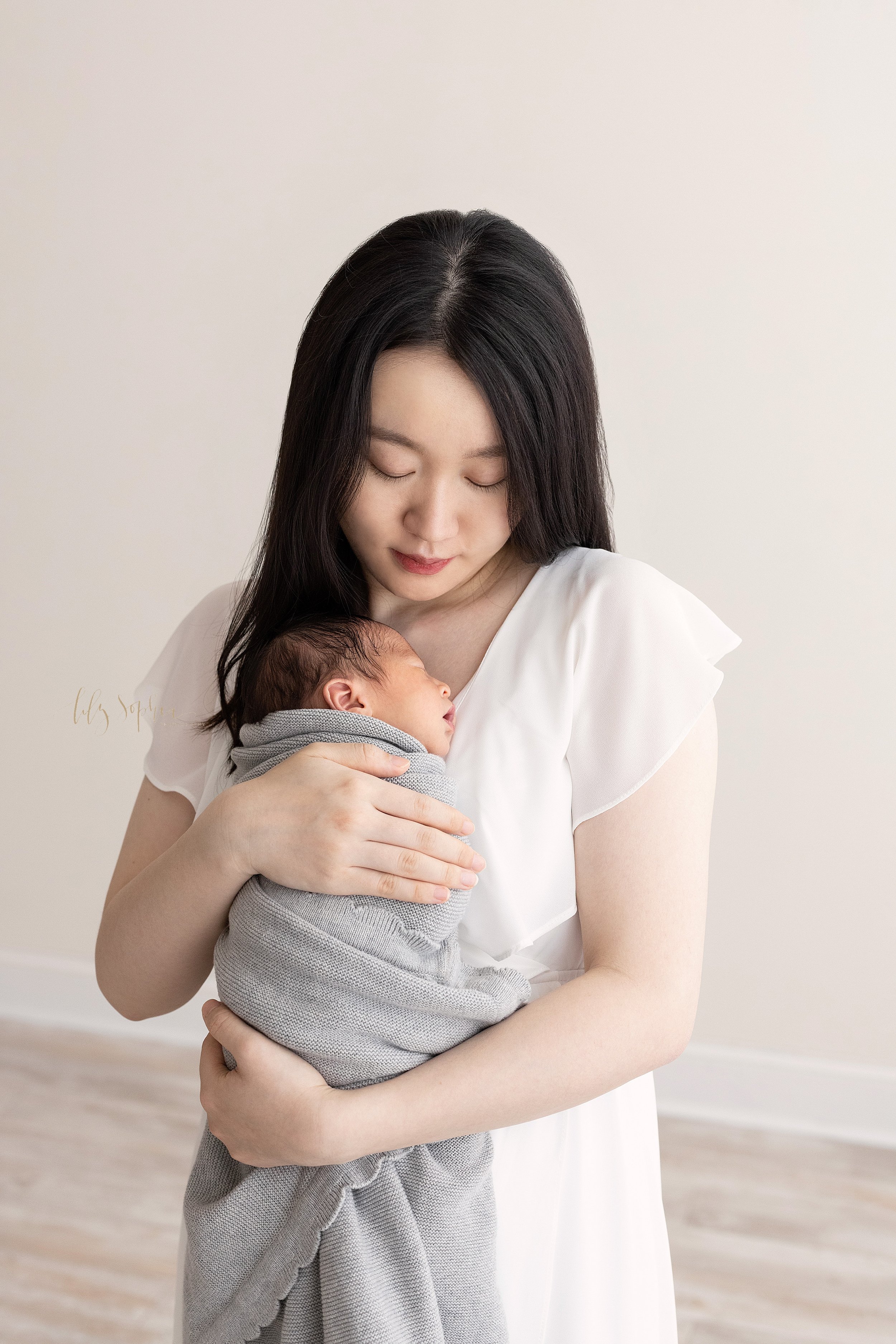  Newborn photo of an Asian mother holding her newborn baby boy against her chest as she stands in a natural light studio near Sandy Springs in Atlanta, Georgia. 