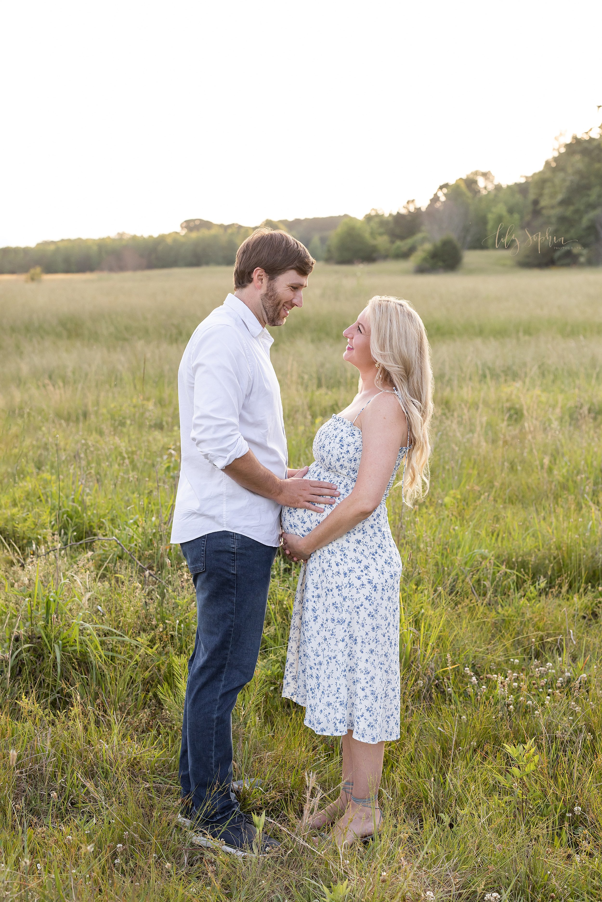  Maternity photoshoot taken in a field near Atlanta at sunset of a husband and wife as they face one another with mom holding the base of her belly and dad placing his hands on their child in utero 