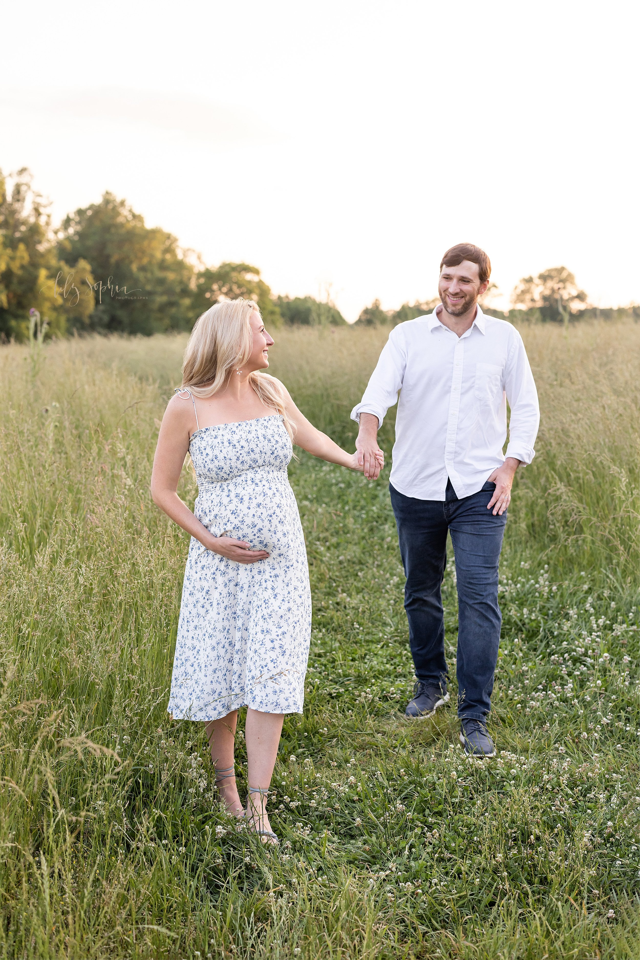  Maternity photoshoot of an expectant couple as they walk hand in hand through an Atlanta field at sunset with the mother holding the base of her belly as she looks over her left shoulder at her husband. 