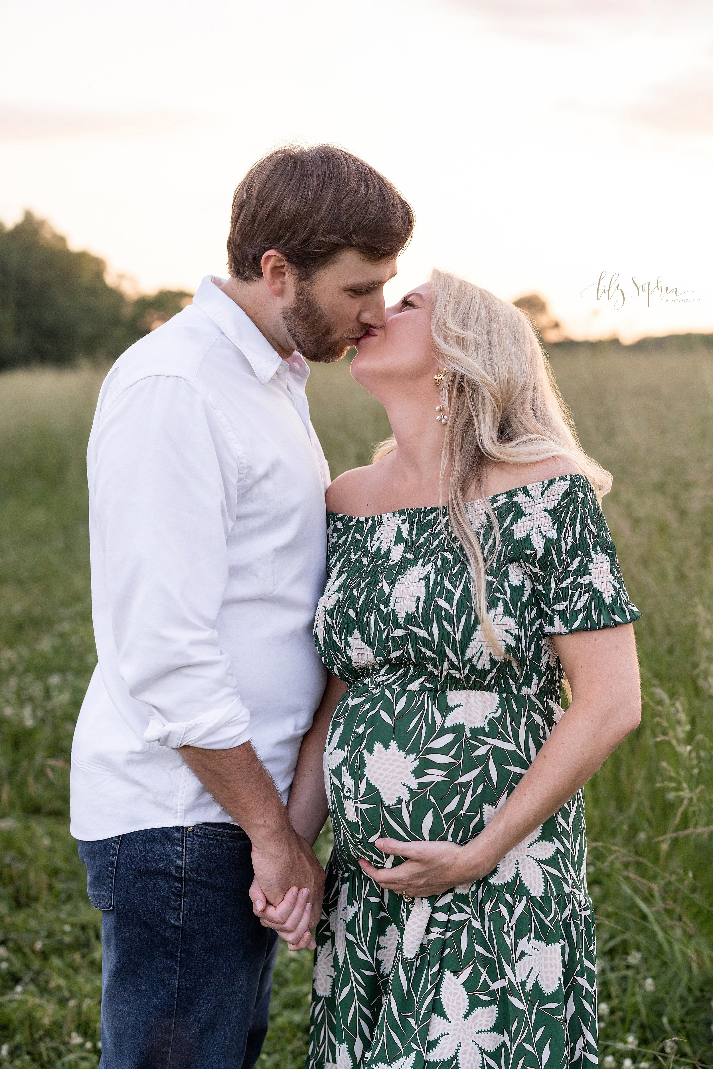  Maternity photo of an expectant mother as she turns to her right to kiss her husband while holding hands with him and holding the base of her belly with her left hand as the couple stand at sunset in a field near Atlanta. 