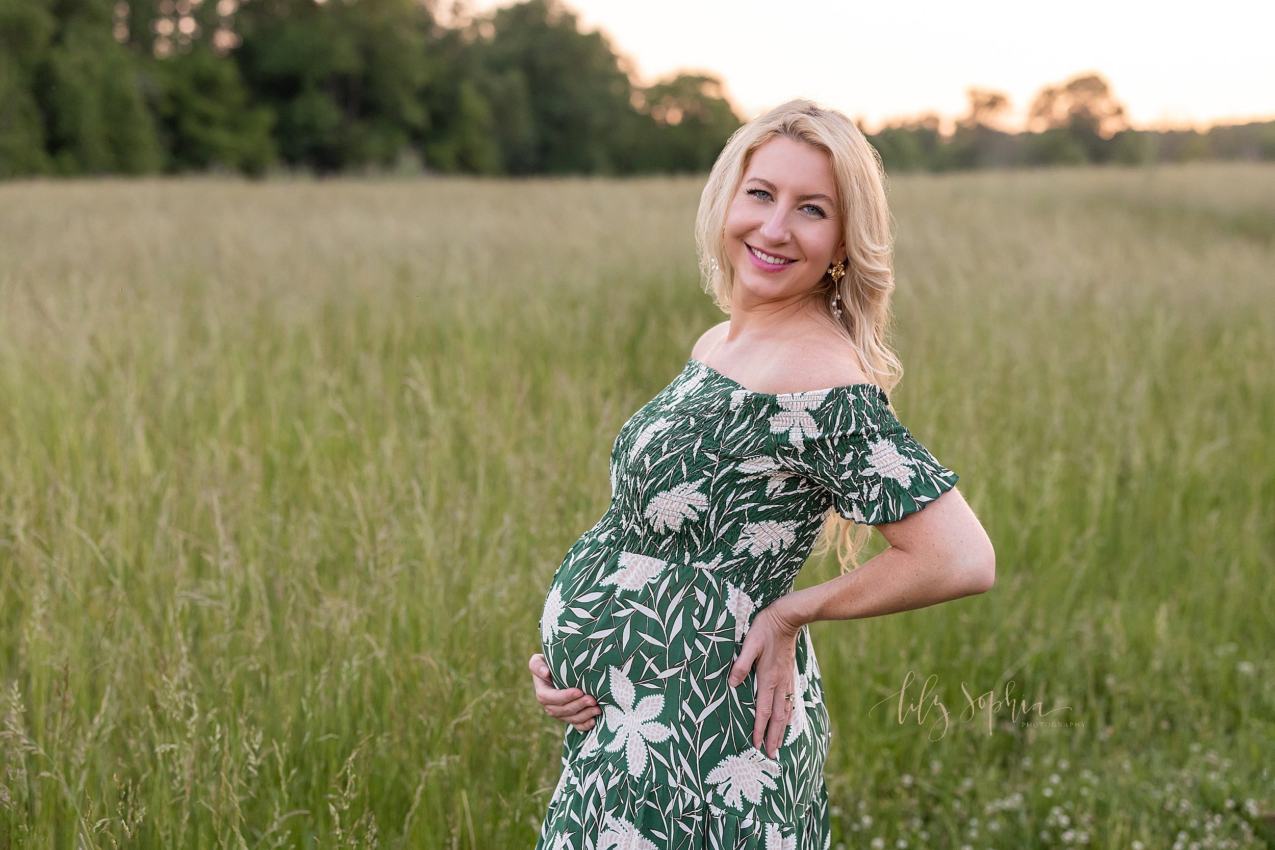  Maternity photoshoot of an expectant mother standing with her right hand at the base of her belly and her left hand on the back of her hip while she smiles and looks over her left shoulder taken in a field at sunset near Atlanta, Georgia. 