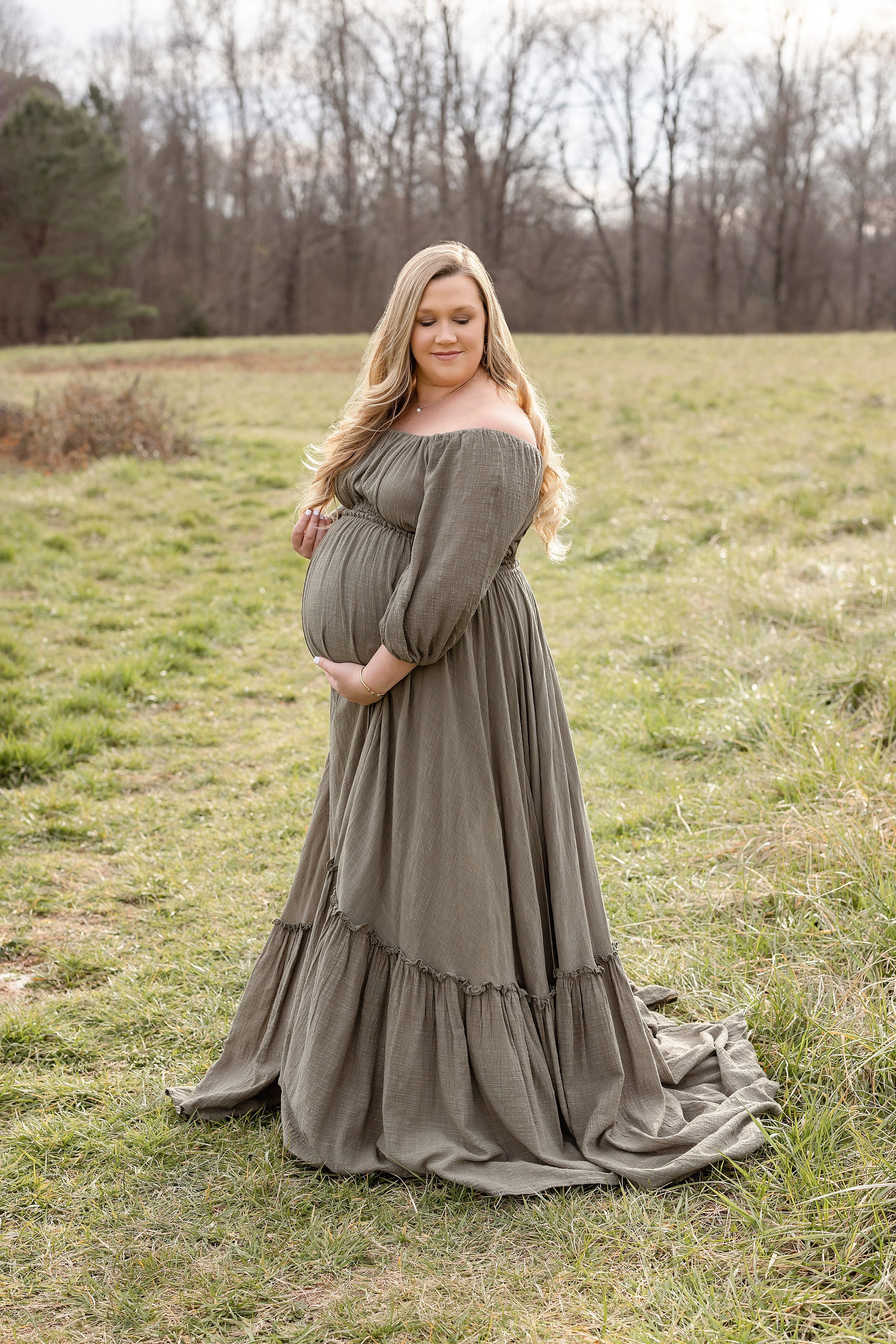  Maternity portrait of a pregnant mother with a peasant off-the-shoulders linen gown standing in an Atlanta field looking over her left shoulder as she plays with her hair with her right hand and frames her belly with her left taken during the fall s