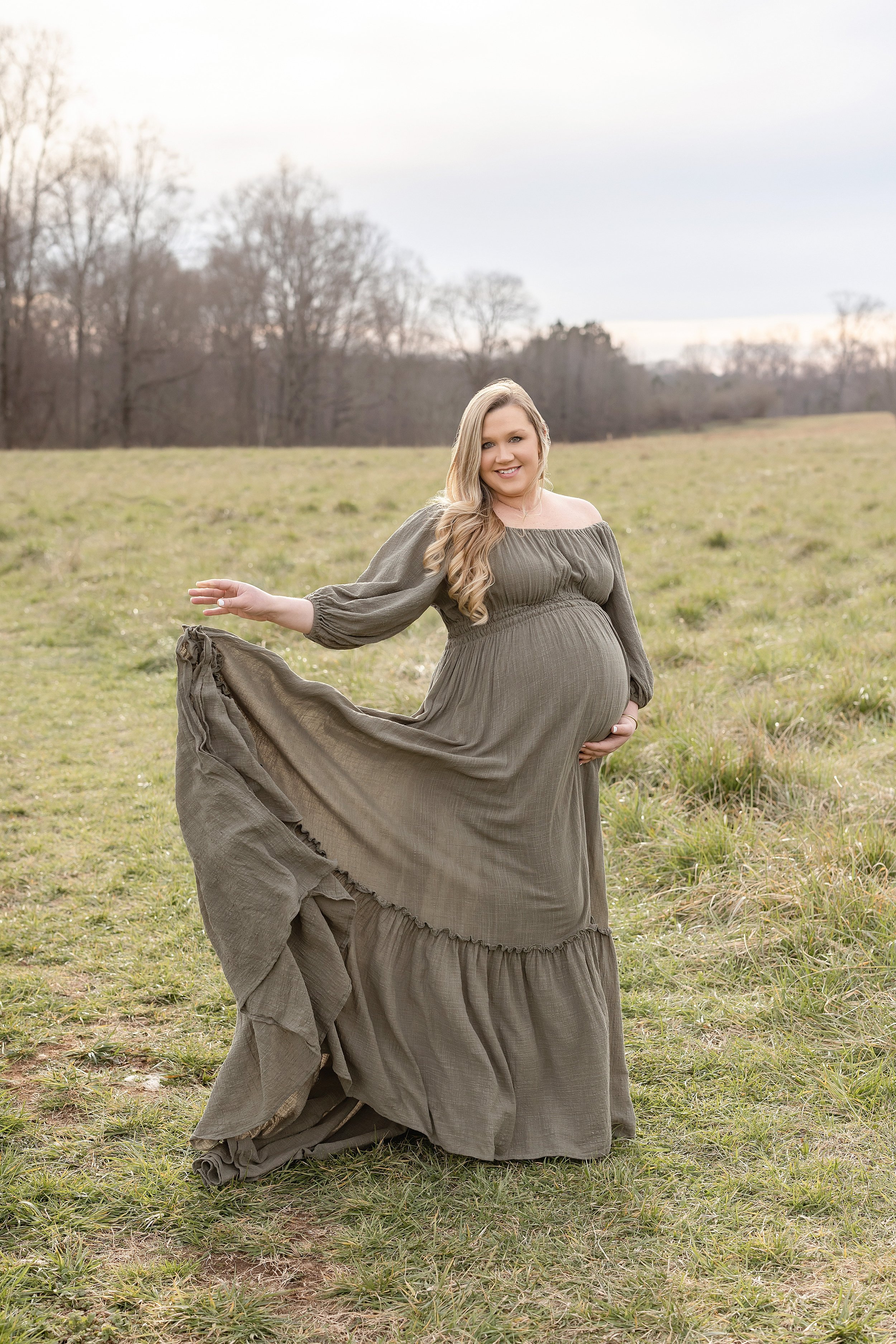  Maternity photo session with a pregnant mother wearing a peasant style linen dress as she stands in a field at sunset with her left hand on the base of her belly and her right hand dropping the hem of her gown taken near Atlanta during autumn. 