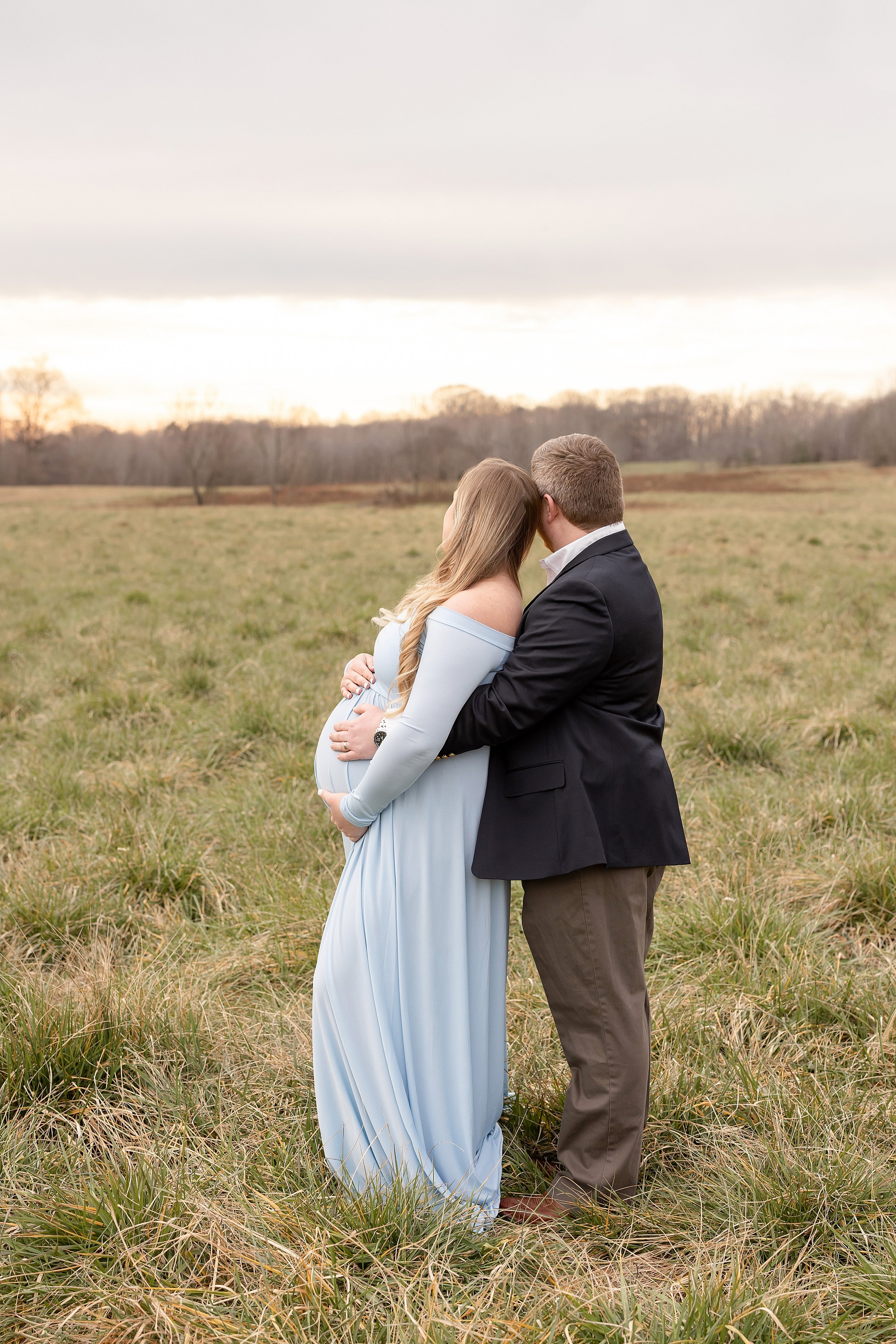  Profile maternity portrait of a pregnant mother framing her belly with her hands as she rests her head against her husband’s head as he stands behind her with his hands laying on her belly and the couple look toward the sunset taken in a field near 