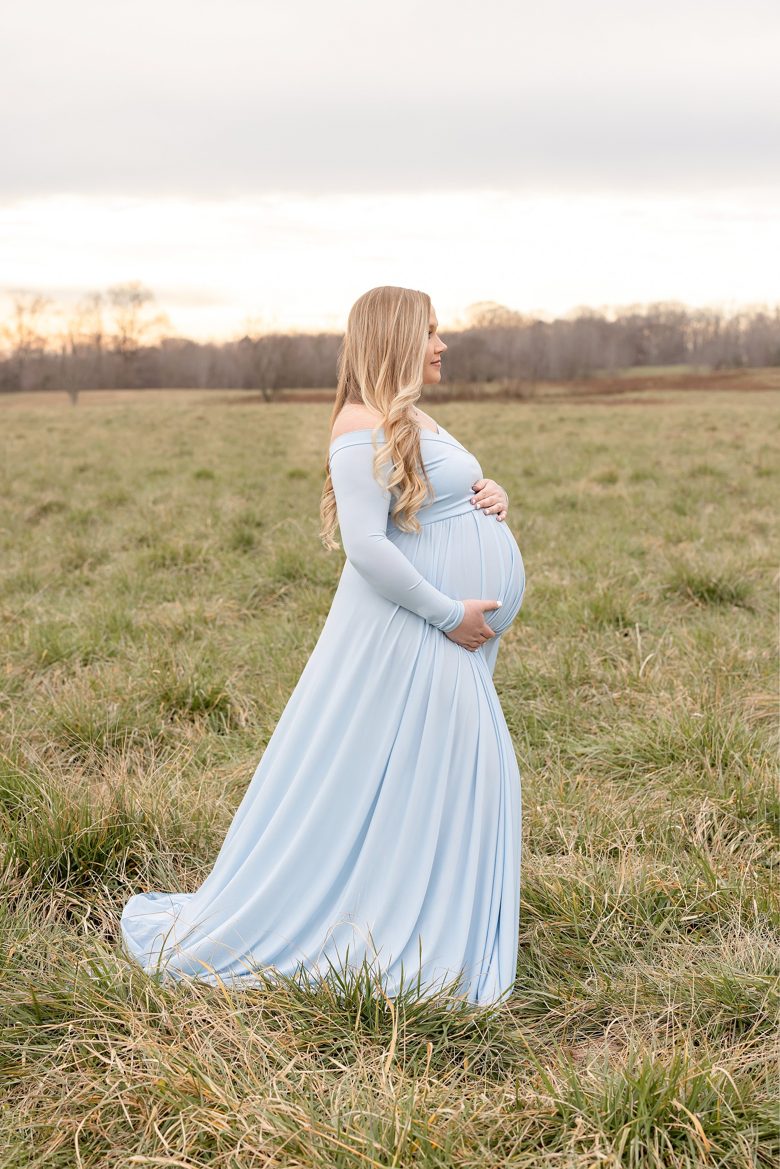  Profile maternity portrait of a pregnant mother framing her belly with her hands as she stands in a field during the fall season wearing an off-the shoulder light blue flowing full-length gown as the sun sets near Atlanta. 