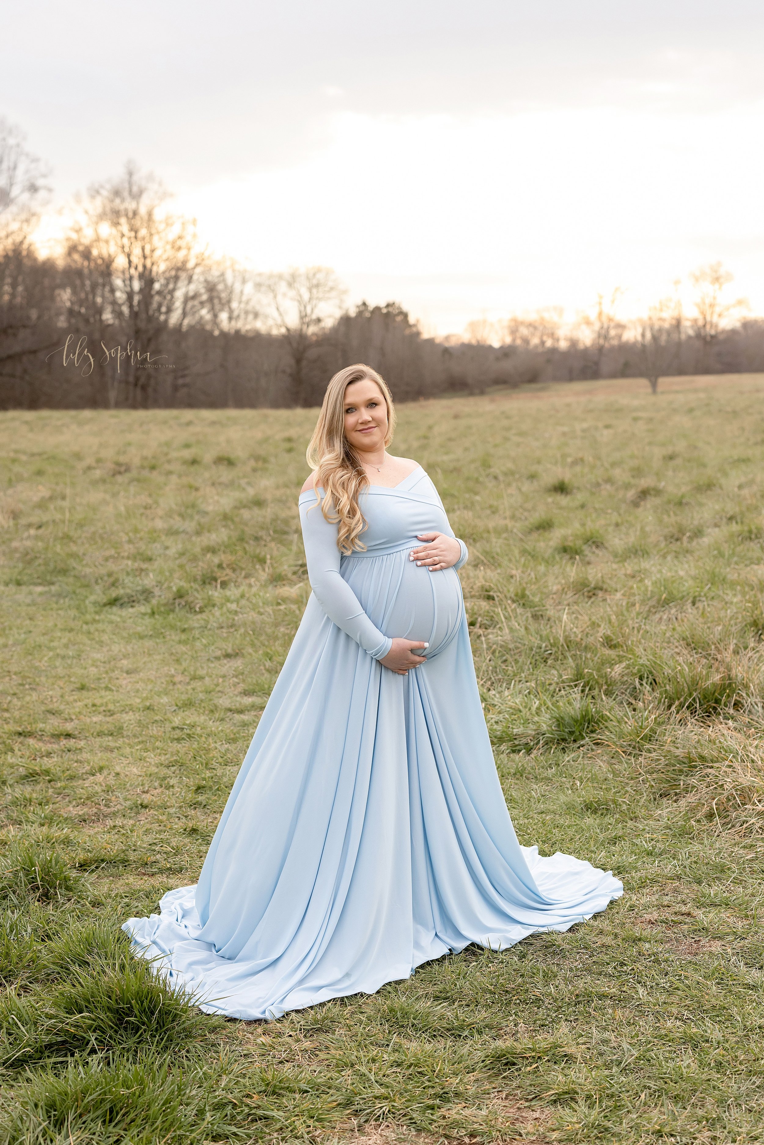  Maternity portrait of a woman standing at sunset in a field wearing a long-sleeve light blue jersey gown who is framing her belly with her hands taken near Atlanta, Georgia. 