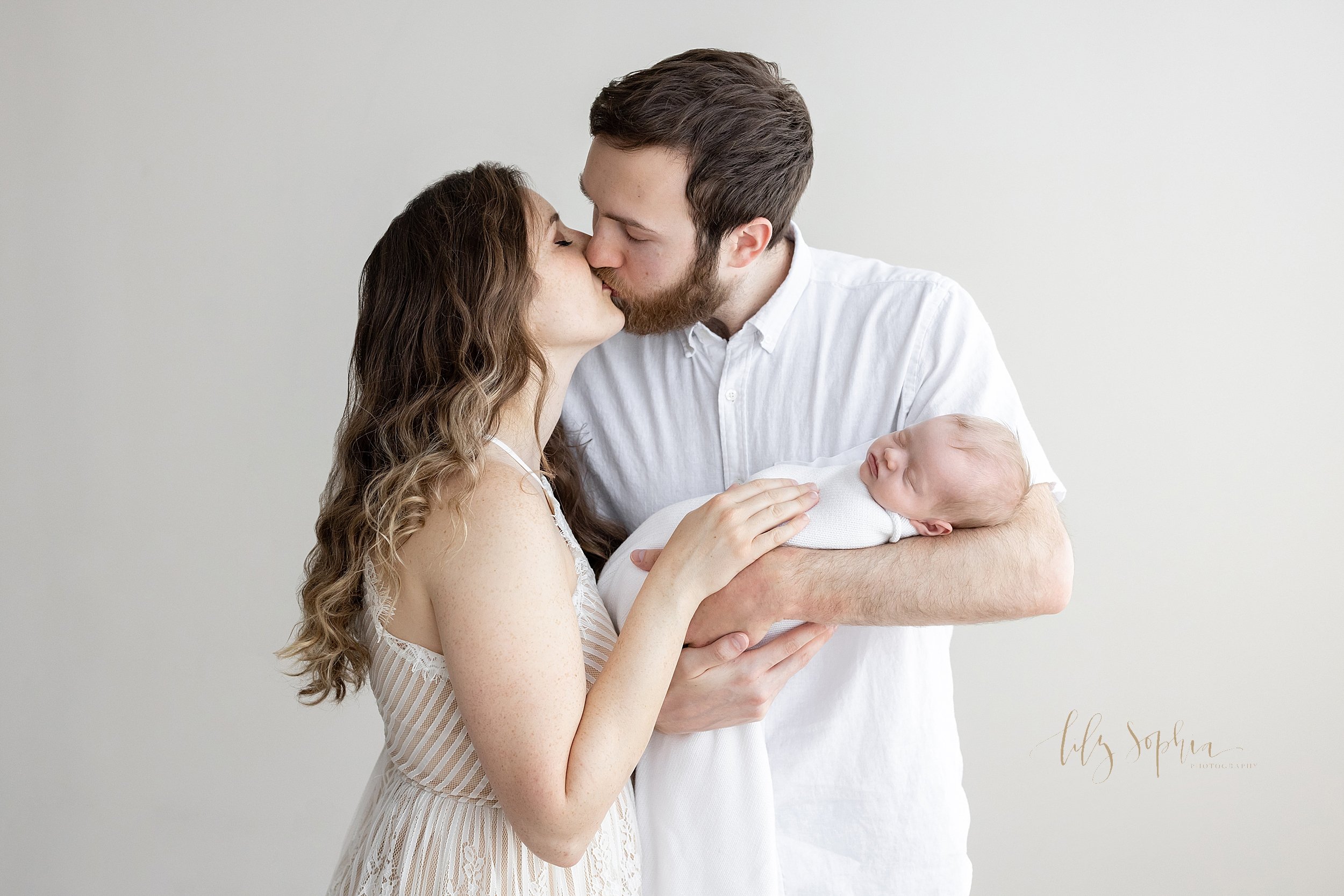 Newborn photo of a father cradling his infant daughter in his arms as his wife stands to his right and the parents kiss taken in natural light near Oakhurst in Atlanta in a photography studio. 