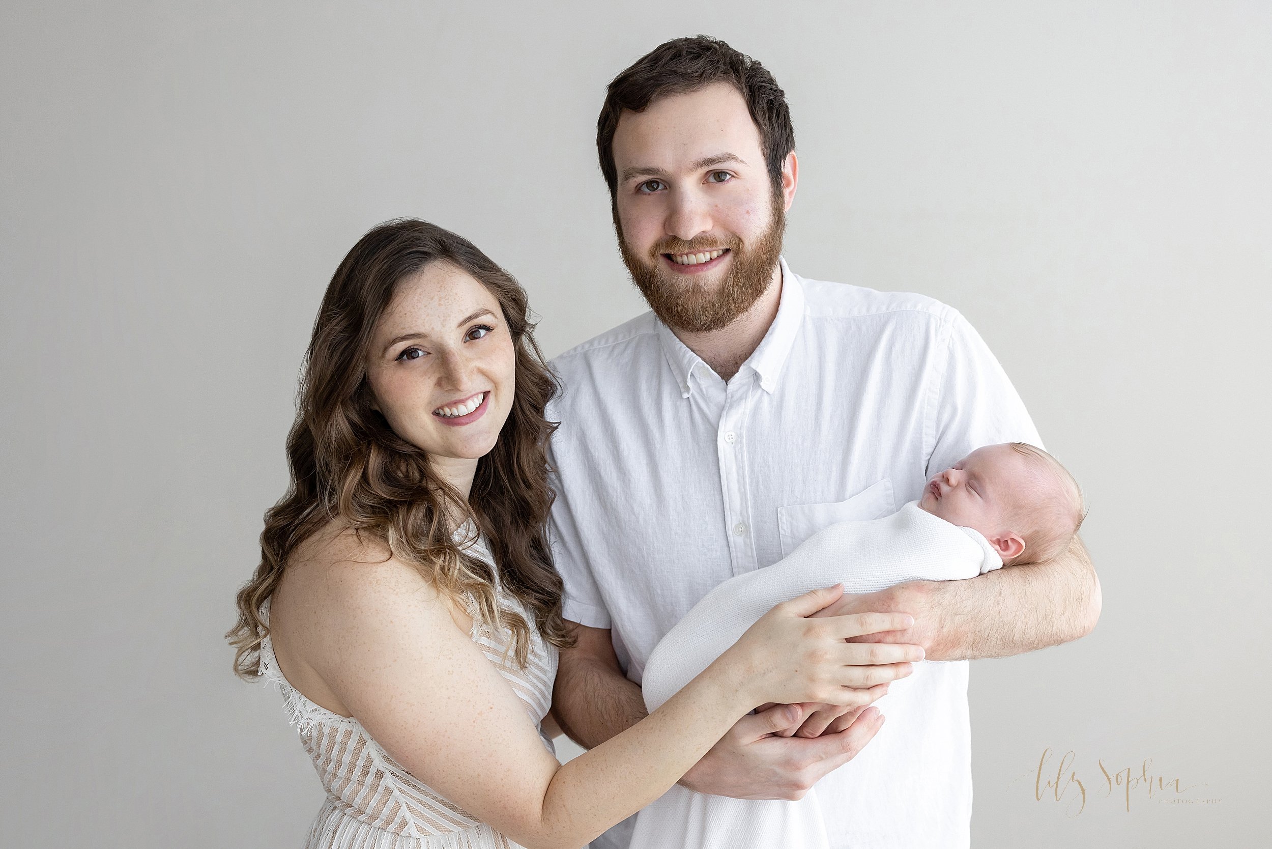  Family newborn photo session with a father cradling his newborn baby girl in his arms as his wife stands to his right side and looks over her right shoulder while placing her right hand on top of her husband’s taken near Old Fourth Ward in Atlanta i