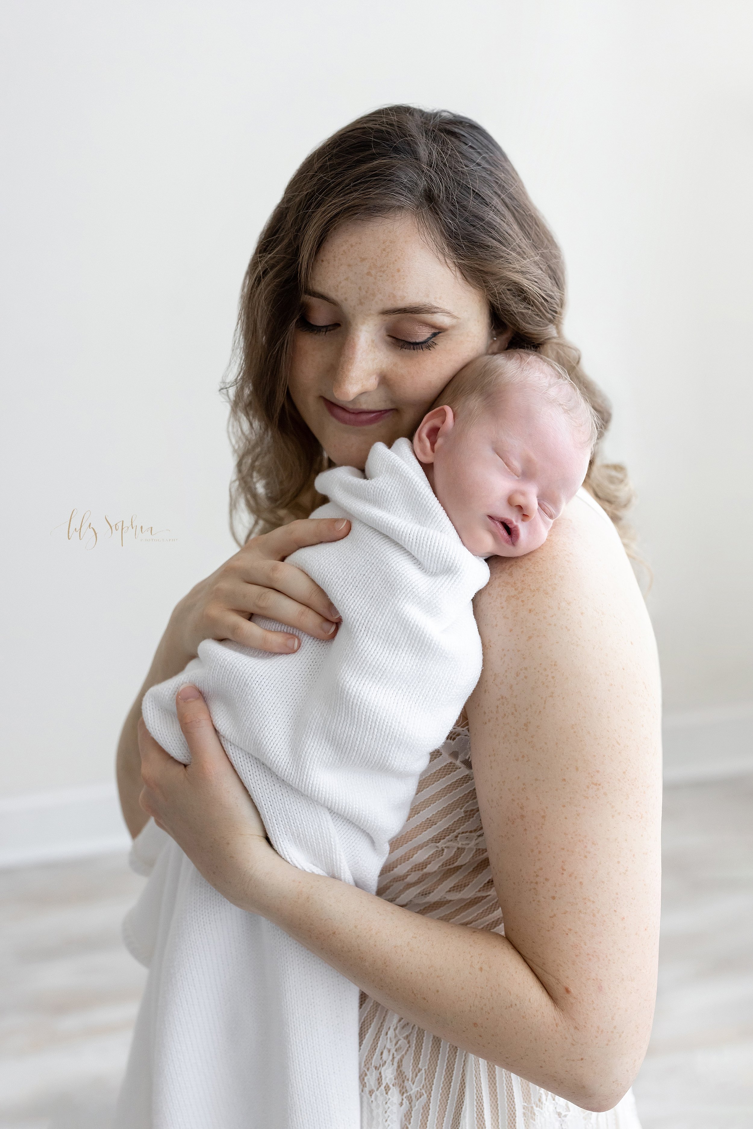  Newborn portrait of a mother holding her sleeping infant daughter on her right shoulder  as she stands in front of a window streaming natural light in a studio near Kirkwood in Atlanta and treasures these moments. 