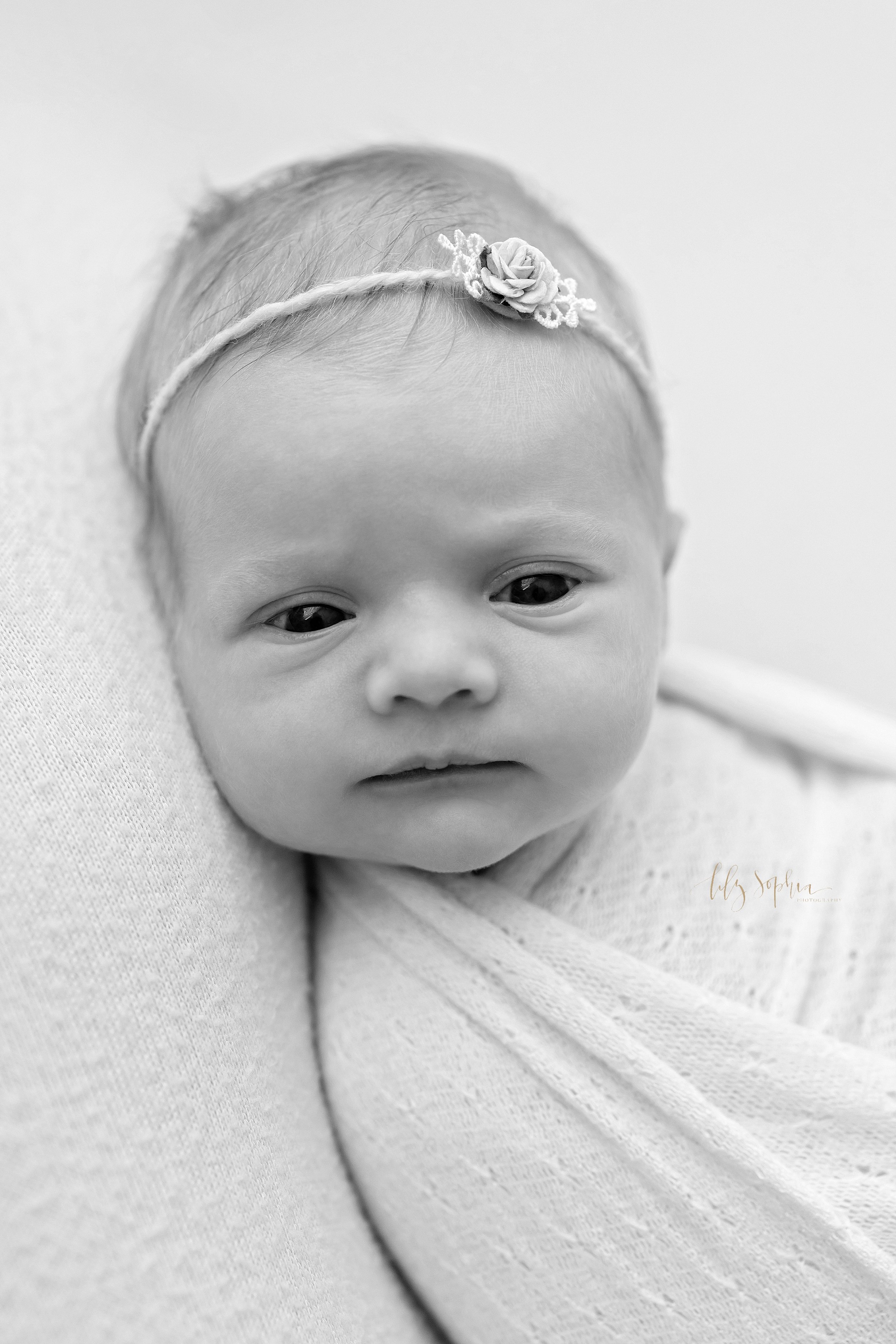  Black and white close-up newborn photo of an awake infant girl wearing a rose headband in her hair taken near Buckhead in Atlanta in a photography studio. 