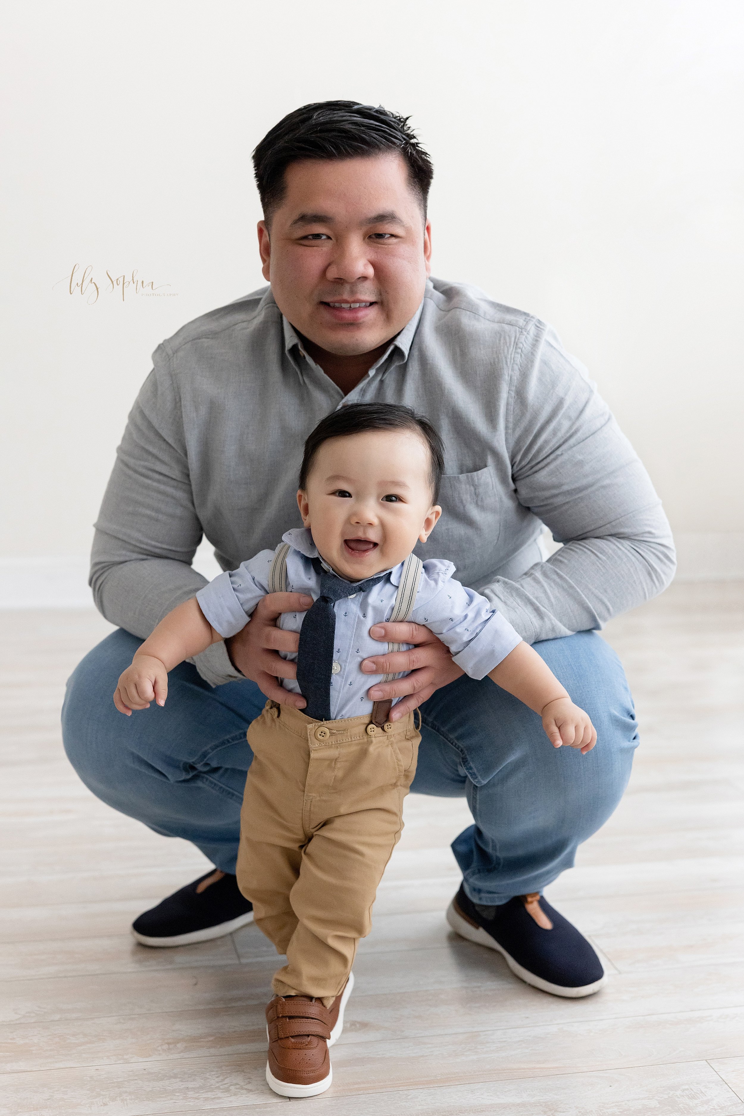  Family photo session of an Asian father squatting behind his baby boy as he supports him to stand taken in front of a natural light window in a studio near Cumming in Atlanta, Georgia. 