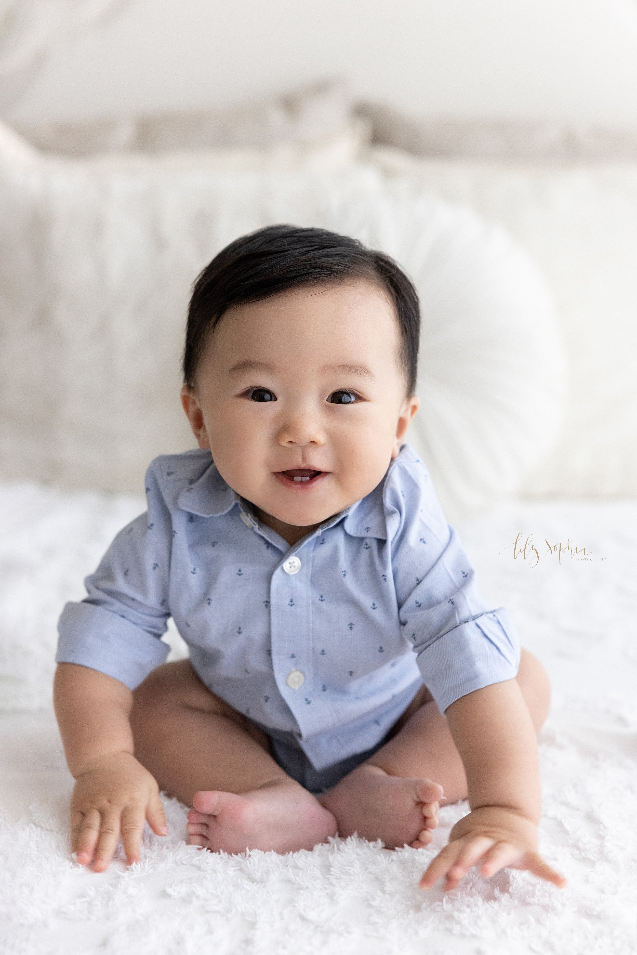  Milestone baby photo of an Asian baby boy as he smiles to show his tiny teeth while sitting on a bed in a studio using natural light in Ponce City Market in Atlanta, Georgia. 
