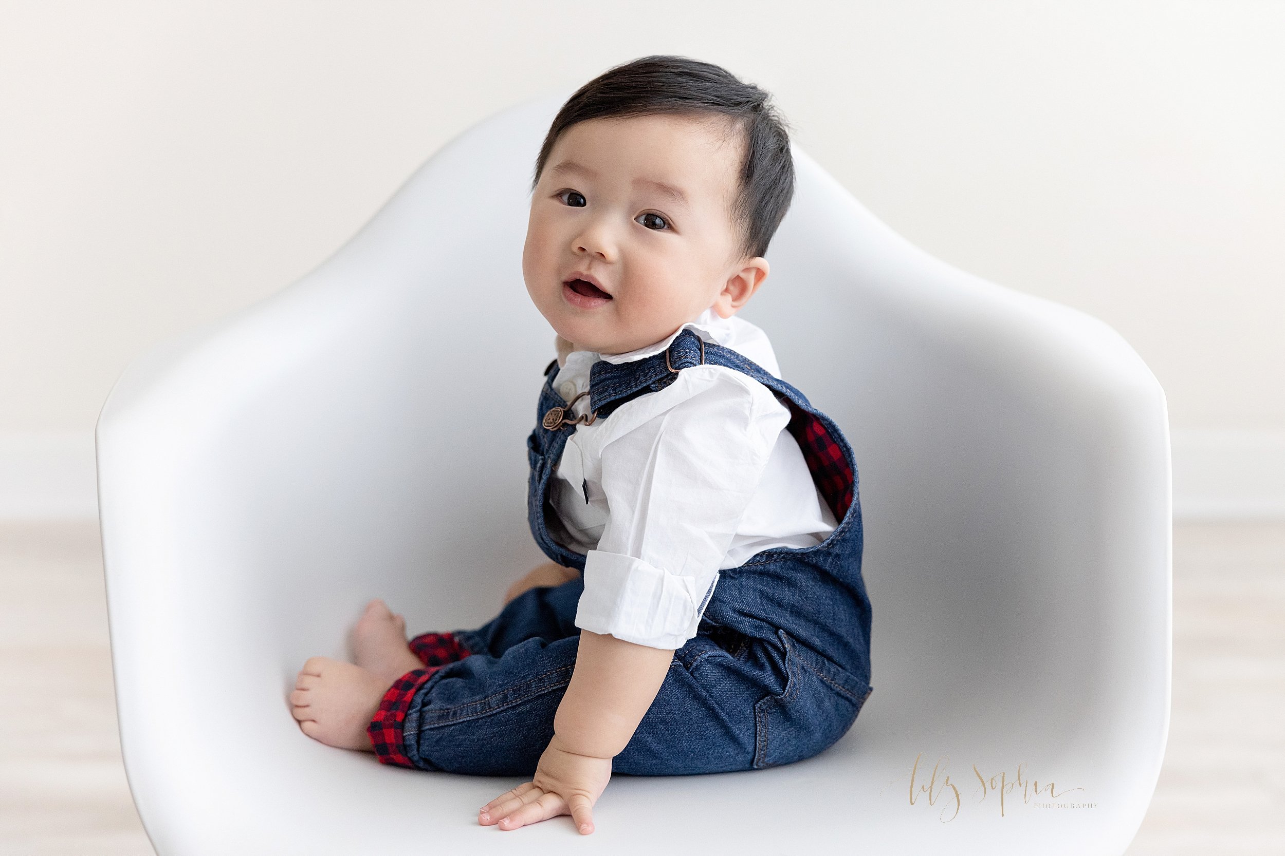  Baby milestone photo of an Asian baby boy wearing denim overalls and a white collared shirt as he sits in a molded white chair and looks over his right shoulder taken in a studio near Sandy Springs in Atlanta that uses natural light. 