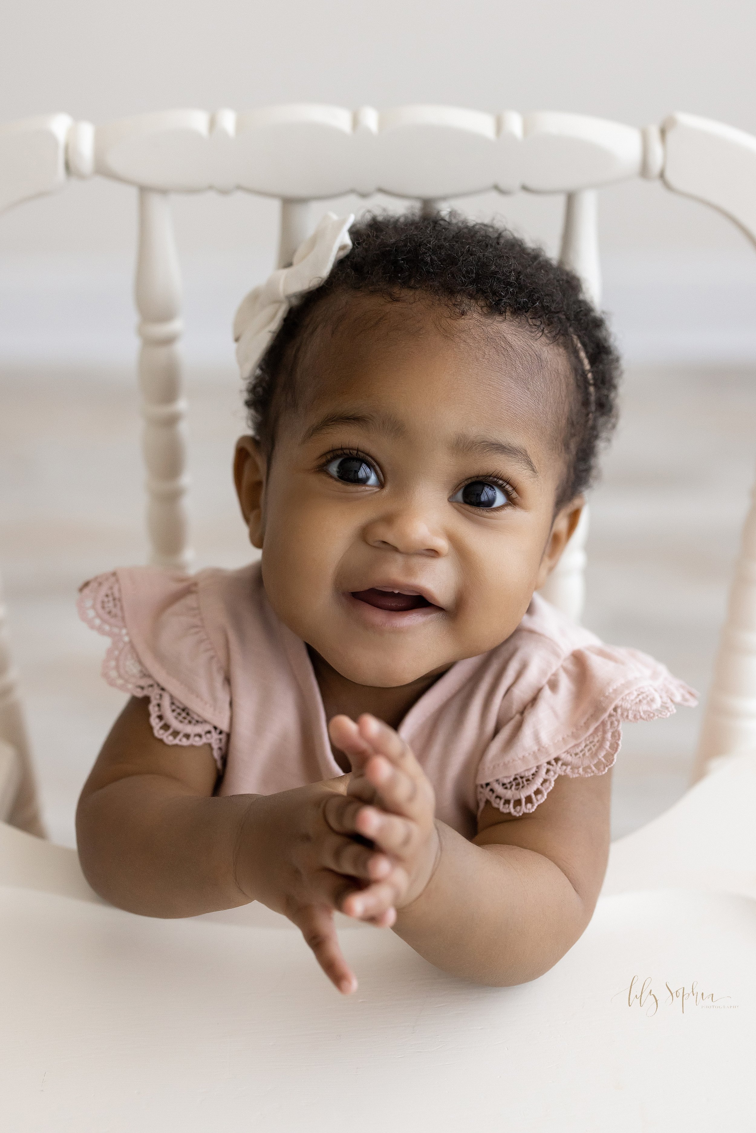  Close-up portrait of an African-American baby girl as she sits in an antique high chair clapping her hands and waiting for her smash cake to celebrate her first birthday taken in a studio near Vinings in Atlanta that uses natural light. 